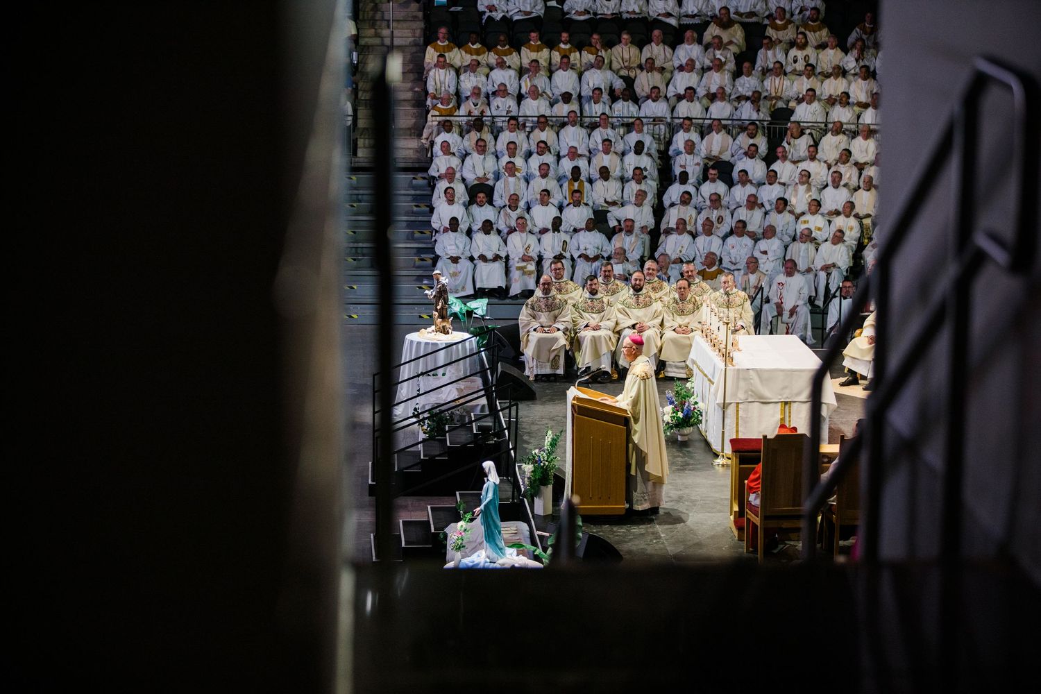 Catholic priests in white vestments celebrate Mass at an altar before a large congregation in an arena.