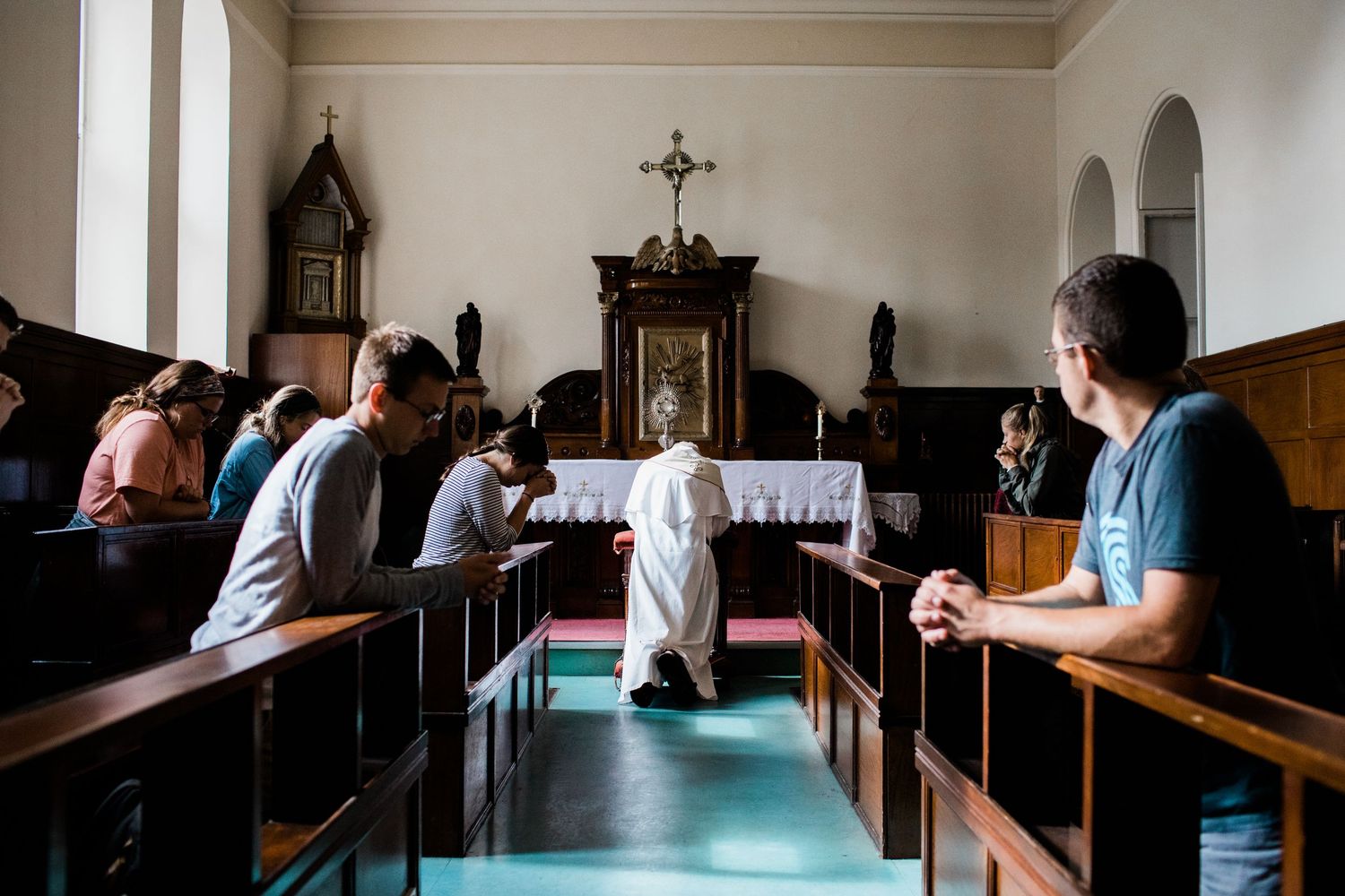 Worshippers kneeling in prayer inside a traditional Catholic church with wooden pews and altar.
