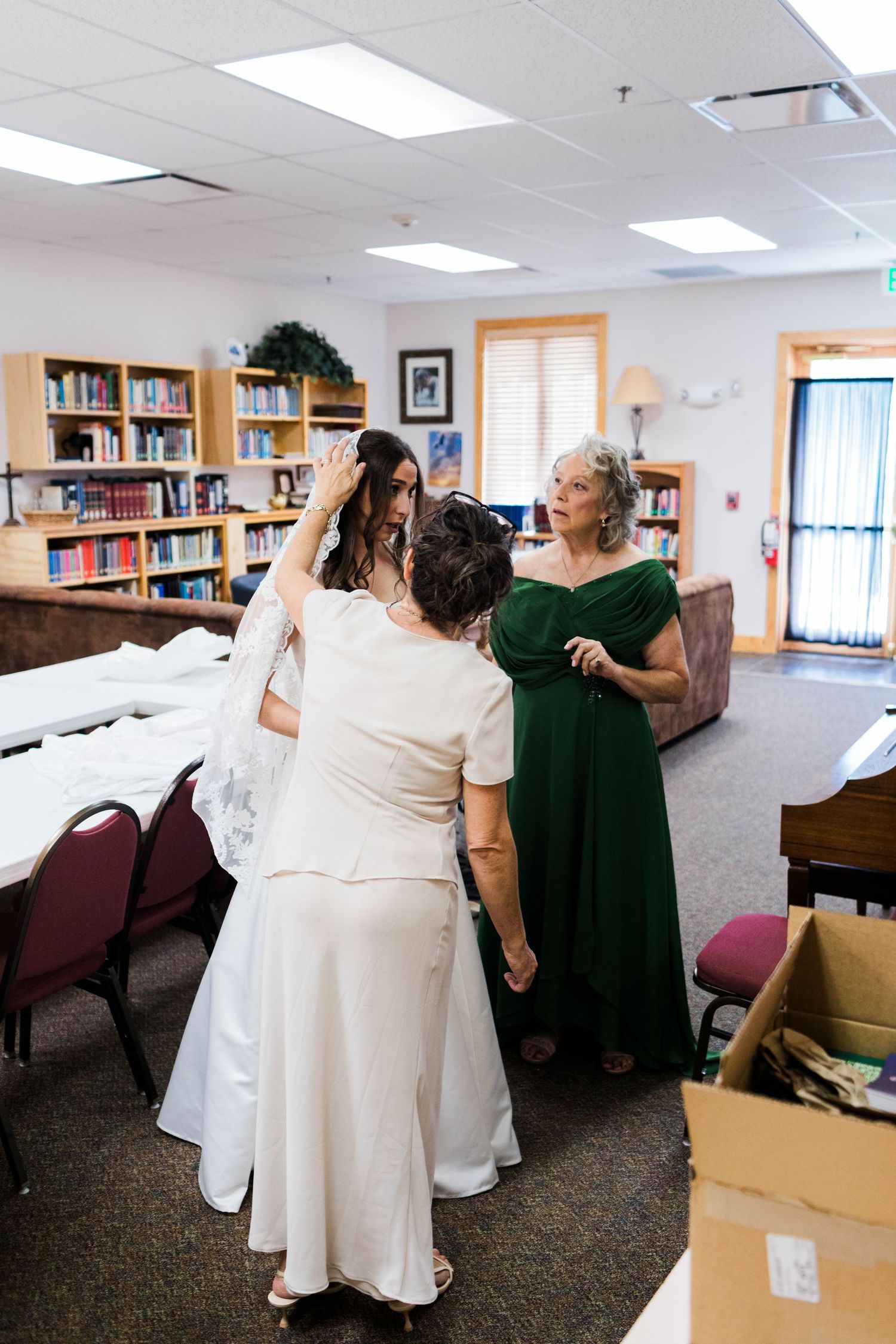 Bride getting veil adjusted by two women in green dress in a cozy library room before wedding ceremony.
