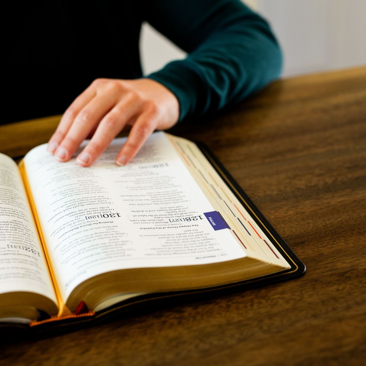 An open Bible resting on a wooden surface with hands visible turning the pages.