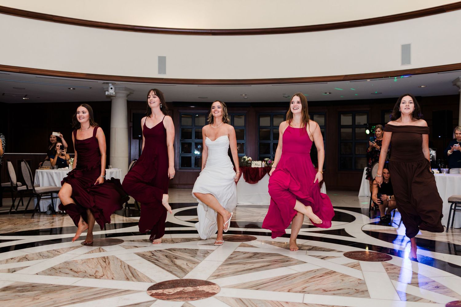 Bride and bridesmaids in colorful gowns dancing joyfully on elegant marble ballroom floor at wedding reception.