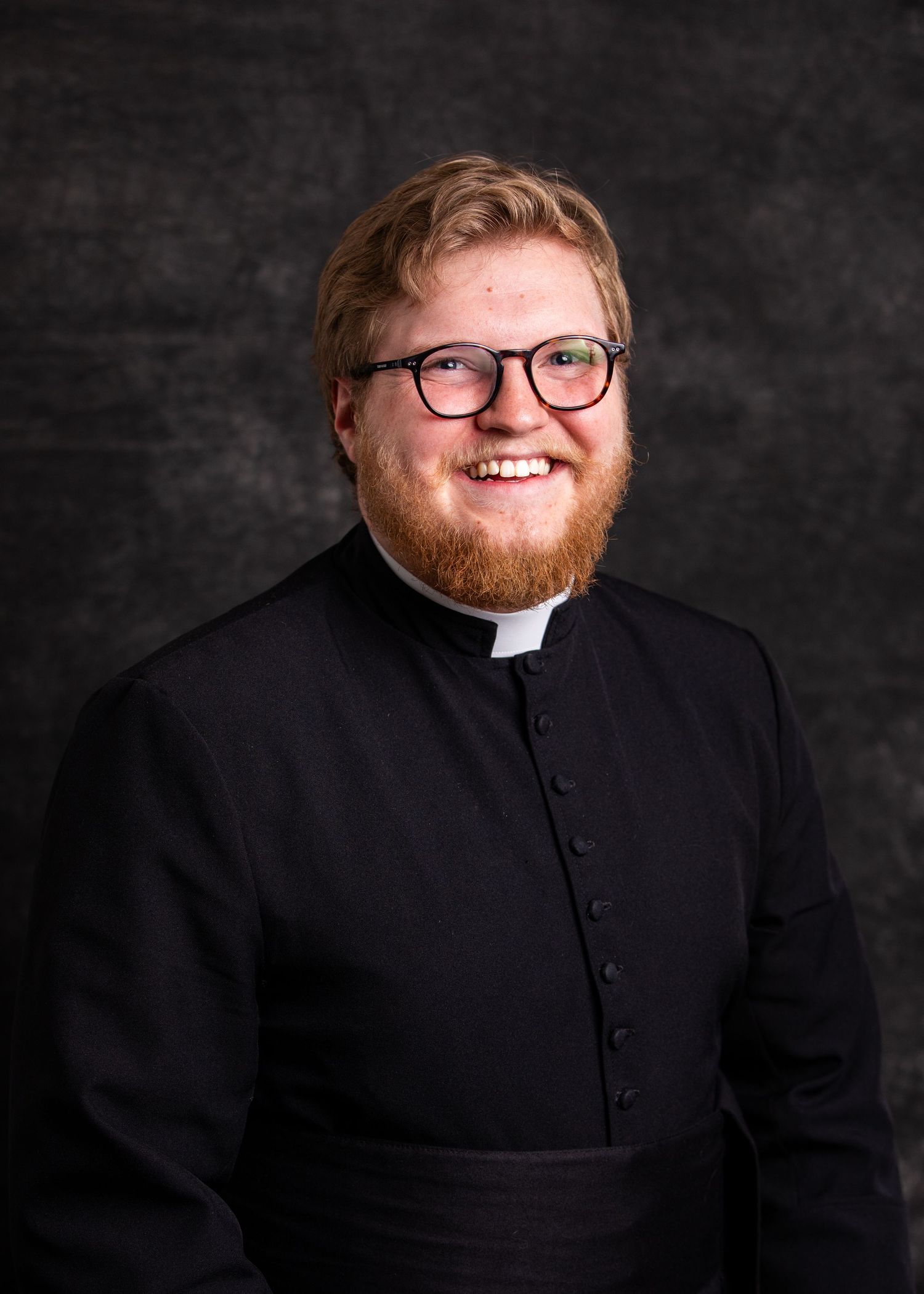 Professional portrait of a clergy member wearing a black clerical shirt and collar against a dark background.