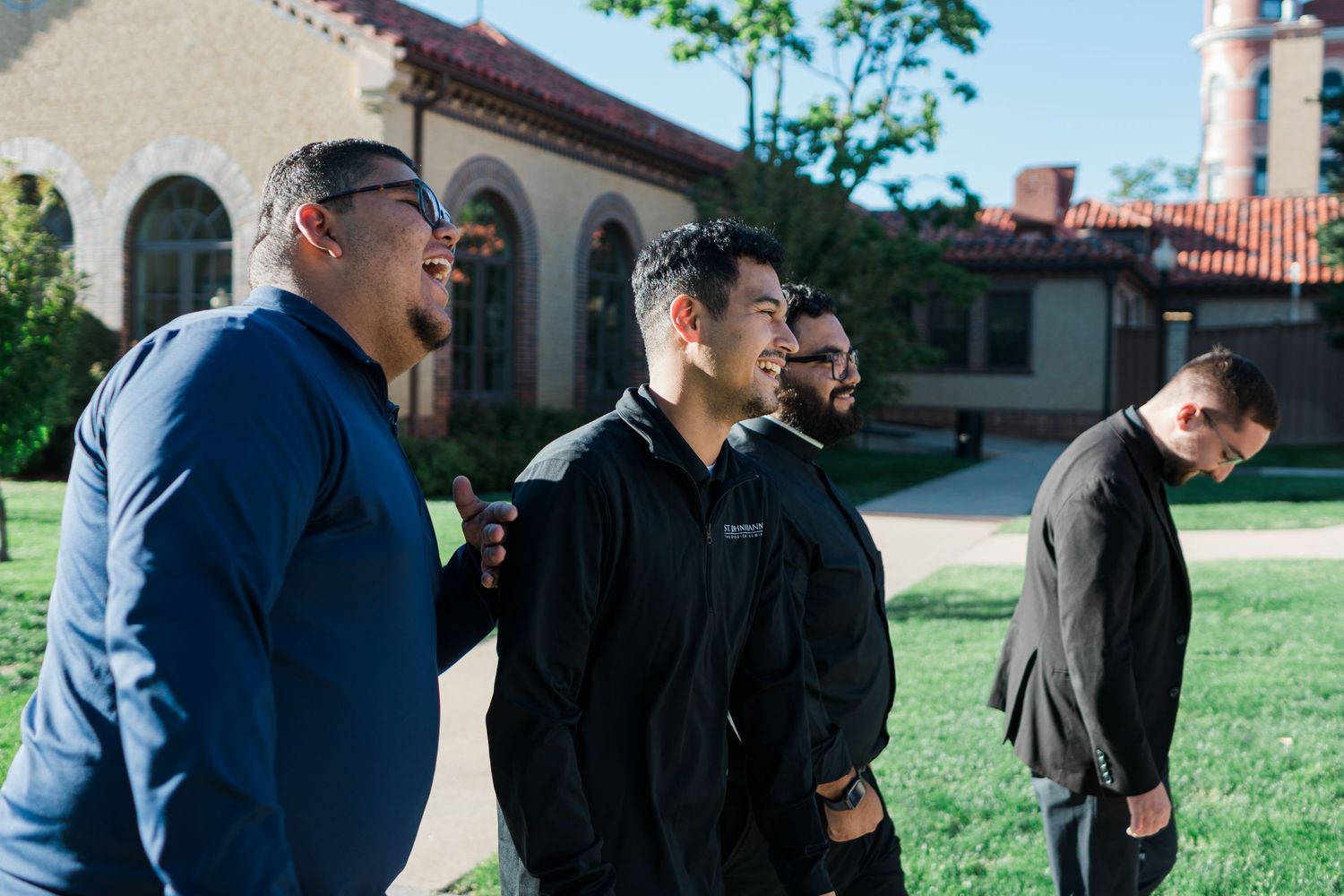 Four young men standing outdoors on a sunny day near a historic brick building with red tile roof.