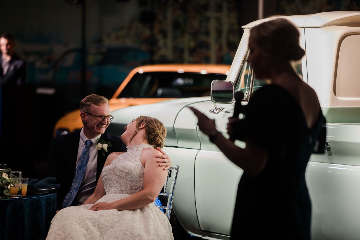 Bride and groom share a kiss beside a vintage car while a guest speaks at their wedding reception.