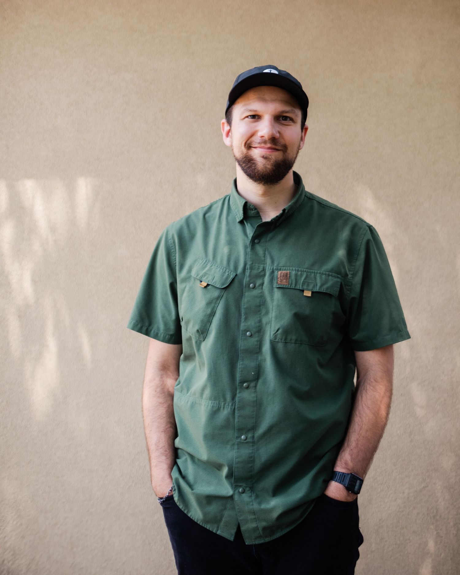 Smiling person wearing baseball cap and green short-sleeved work shirt stands against beige wall.