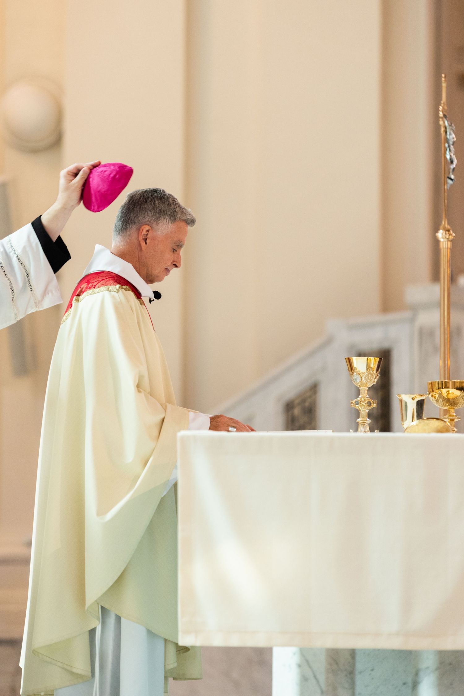 A priest in white robes stands at an altar with a chalice and candles during a religious ceremony.
