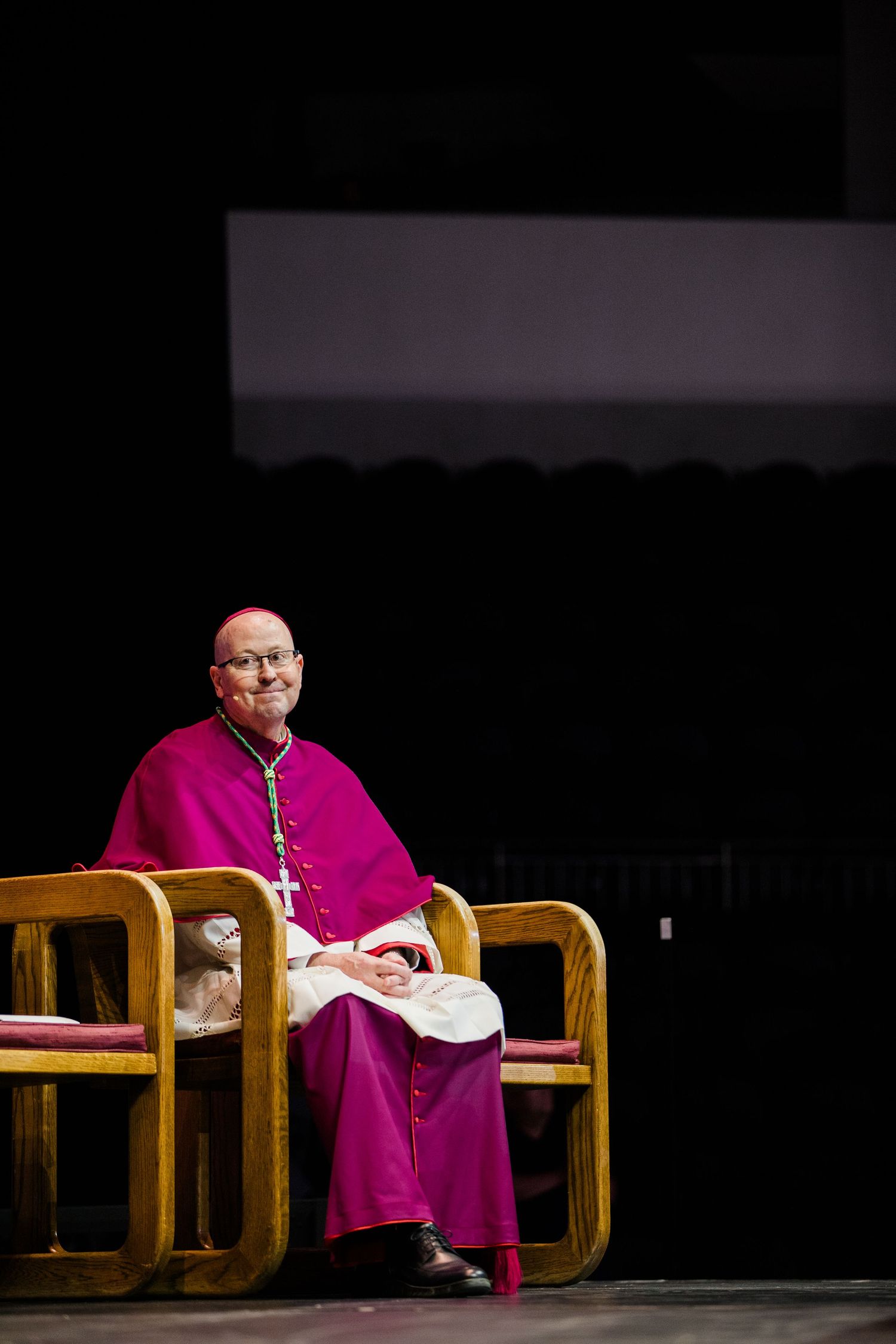 A Catholic bishop wearing a magenta robe sits on a wooden chair in a dimly lit setting.