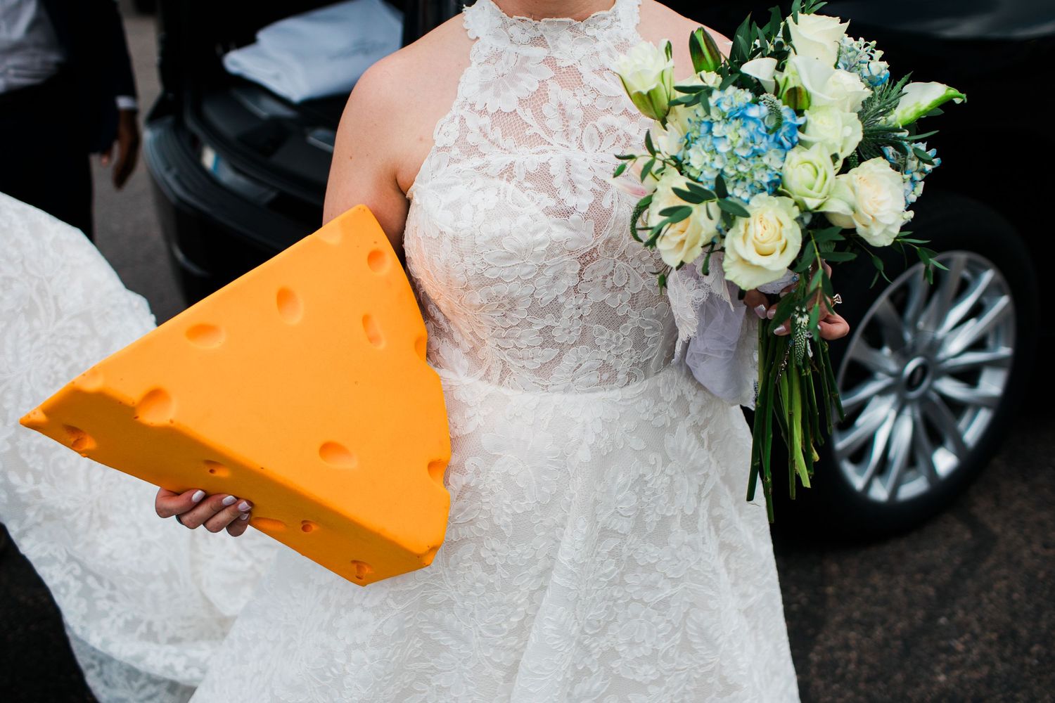Bride in lace wedding dress holding a giant cheese wedge prop and a blue and white floral bouquet.