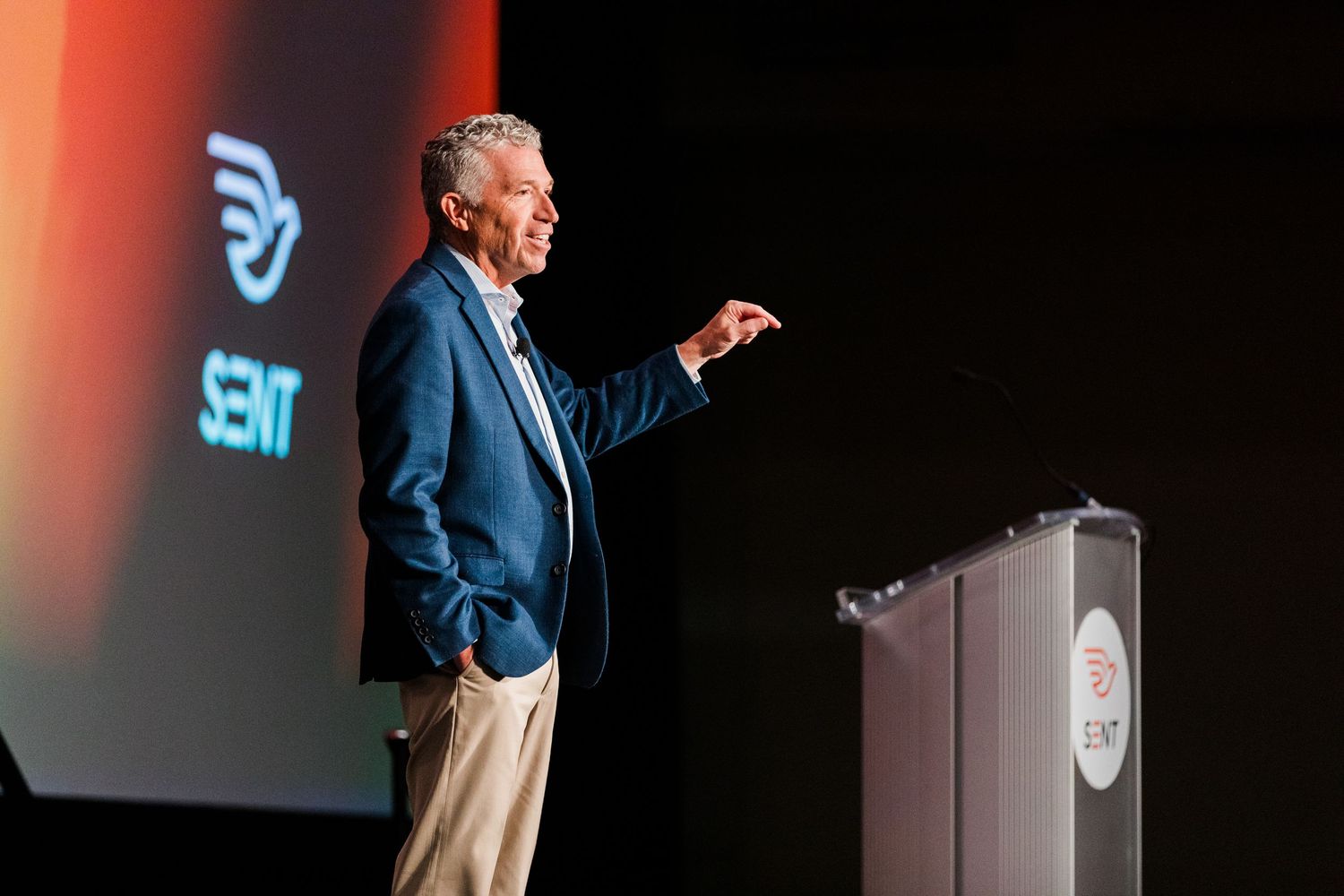 Man in blue blazer speaking on stage at SENT conference, gesturing while presenting at podium.