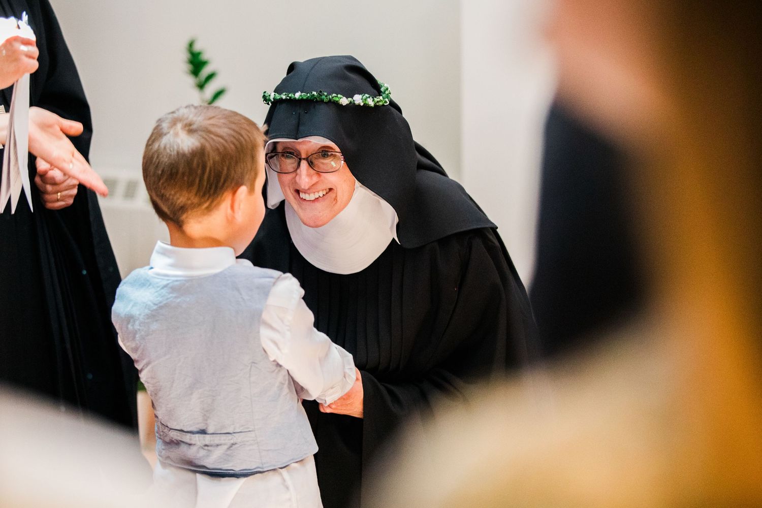 A smiling nun in black habit warmly greets a young child in a white outfit at a religious event.