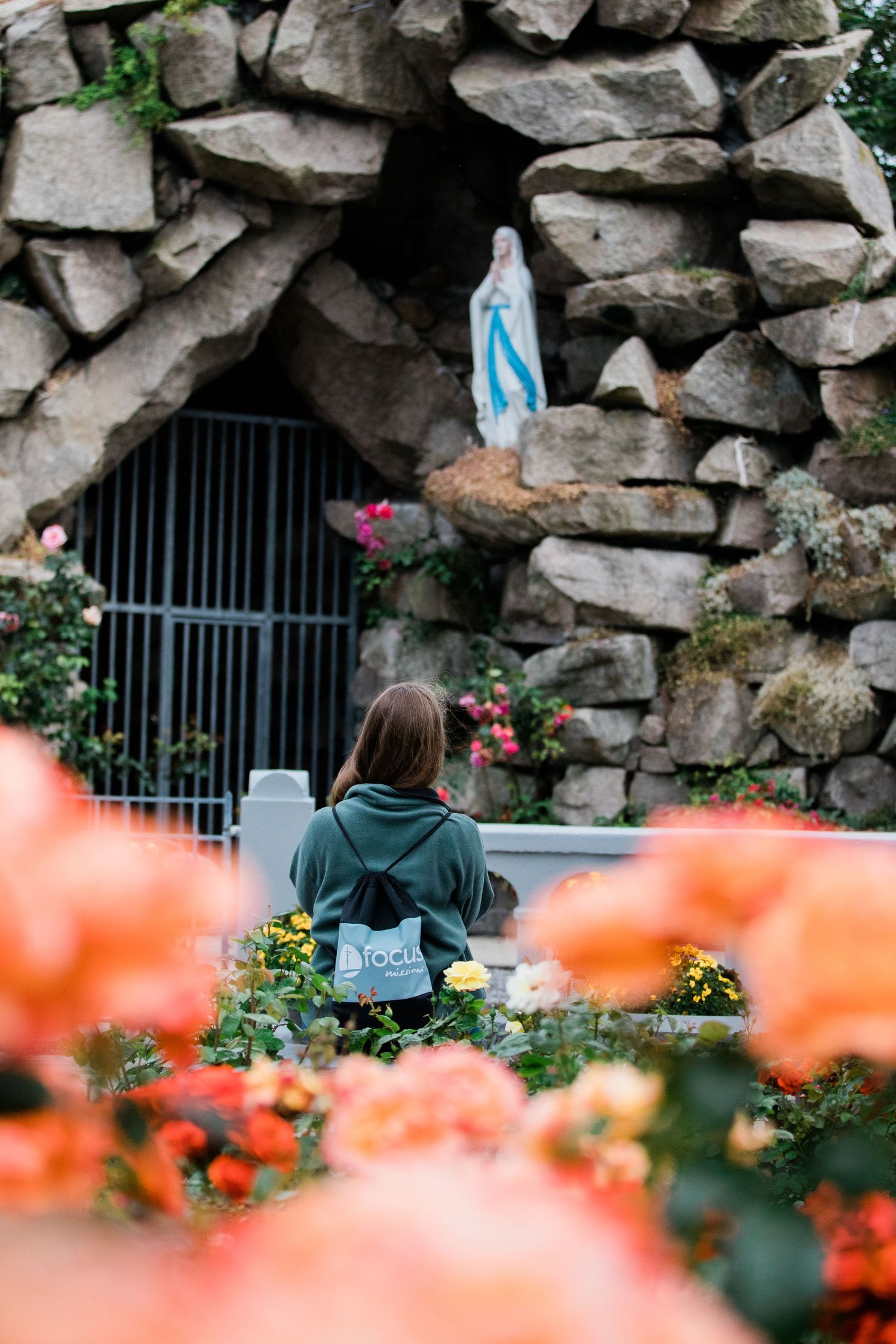 A woman prays before a Virgin Mary grotto shrine surrounded by colorful blooming flowers.