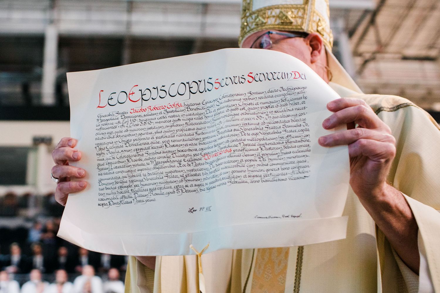 A bishop in golden vestments holds up an ornate illuminated papal document during a church ceremony.