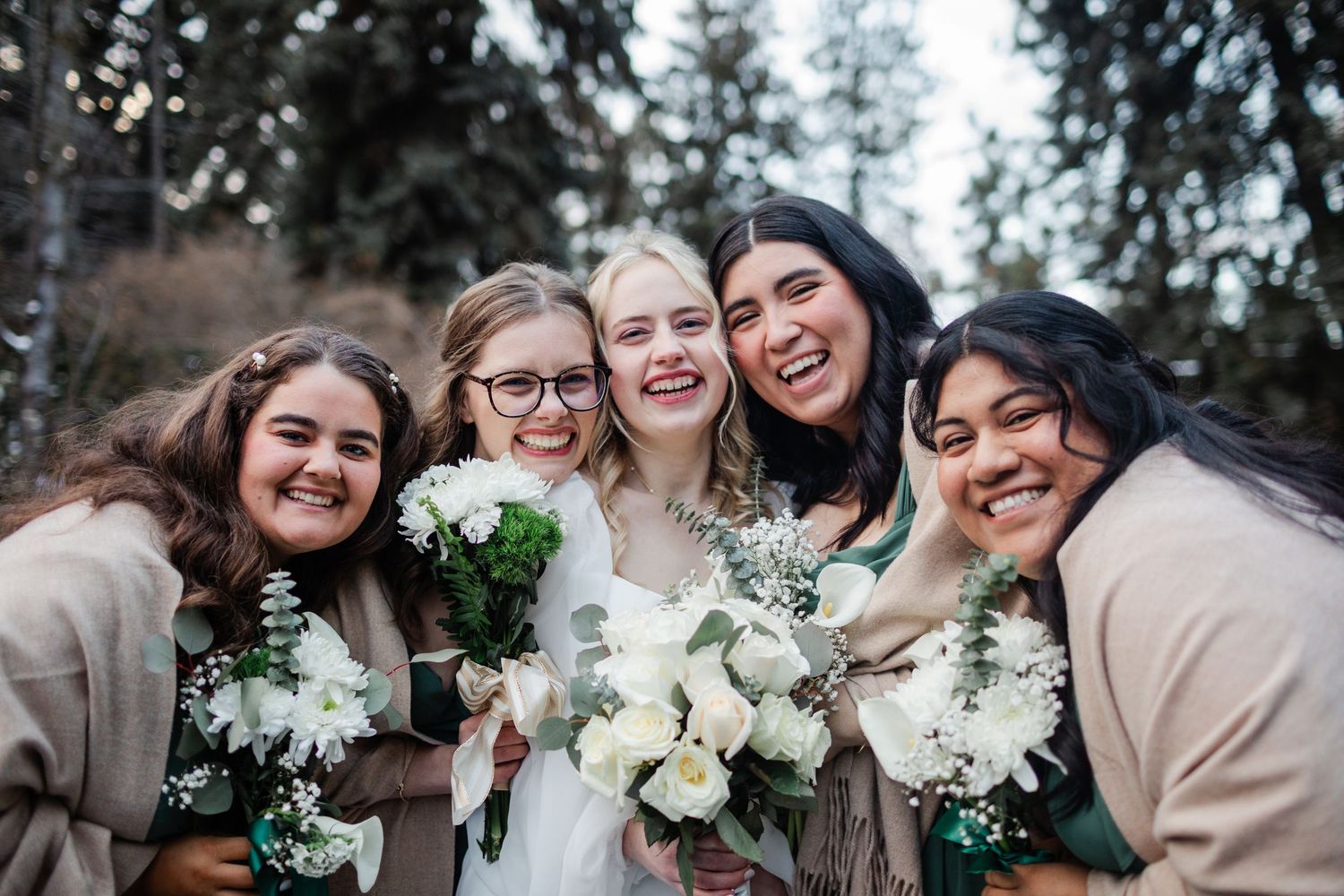 A group of bridesmaids wearing neutral-colored dresses pose together while holding white floral bouquets.