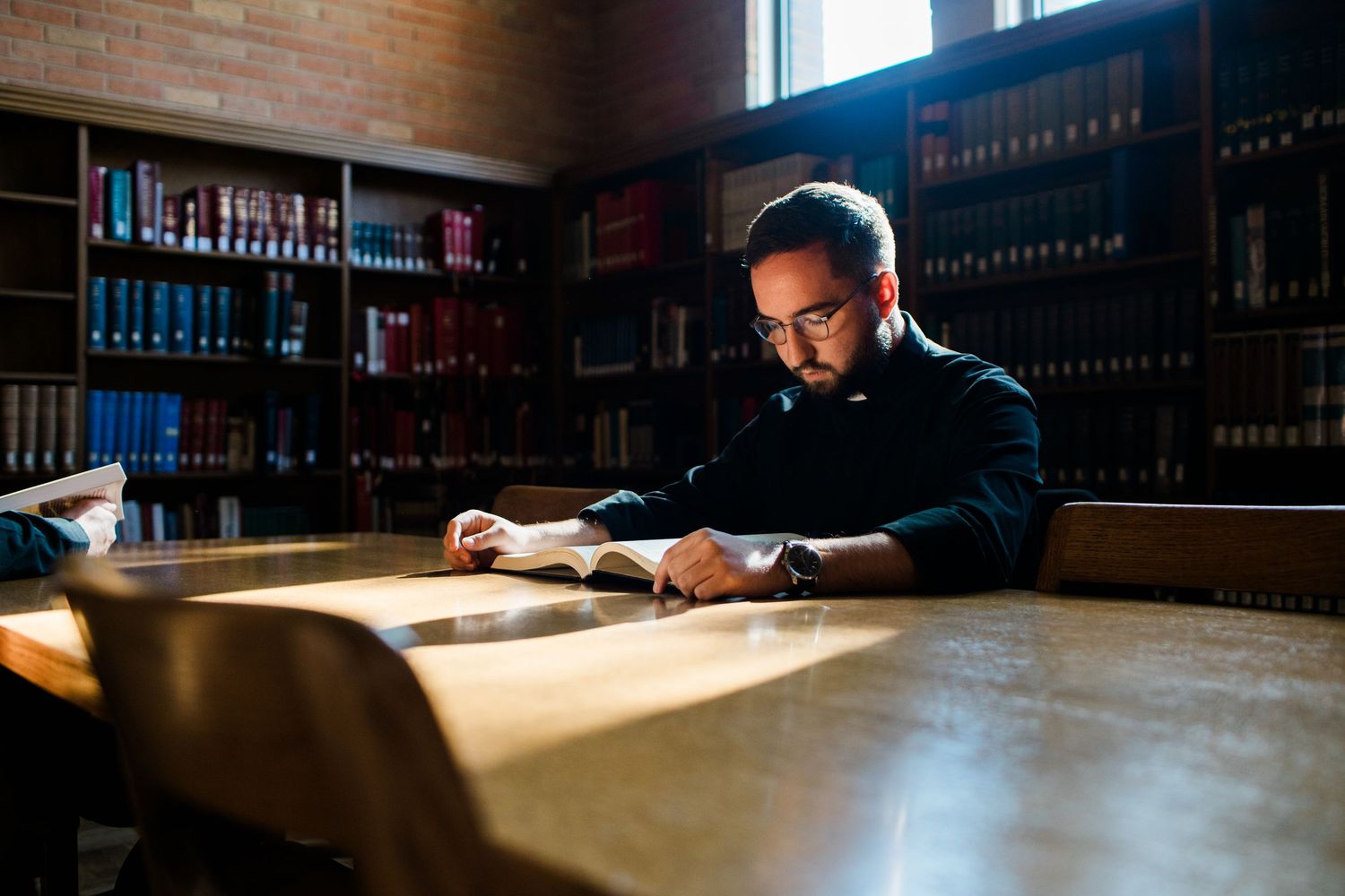 Man with glasses reading a book at a wooden table in a dimly lit library with bookshelves in the background.