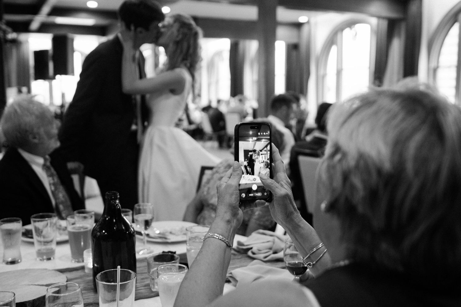 Black and white photo of wedding guests photographing the bride and groom sharing a first dance at their reception.