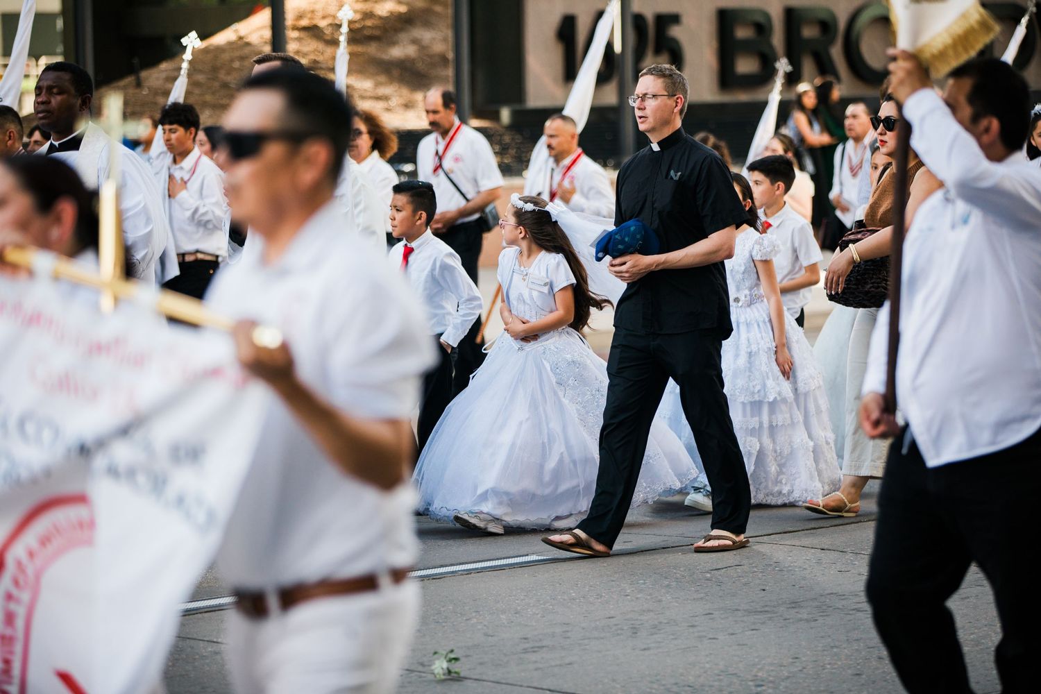 A festive street parade featuring performers in white dresses and formal attire marching along a city boulevard.