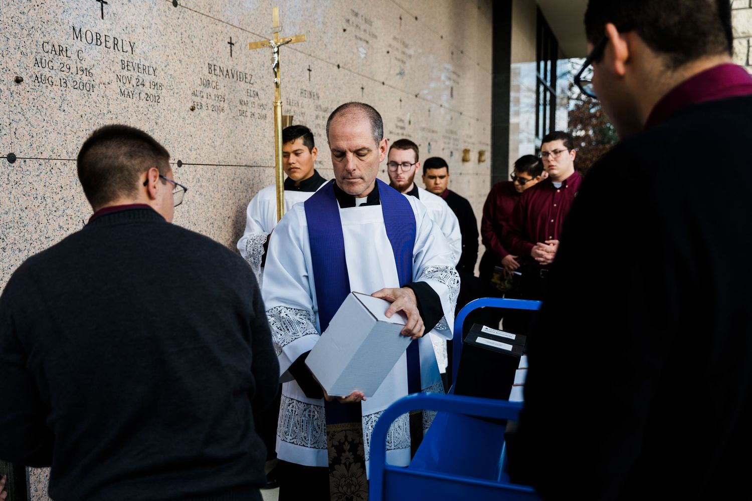 A person in a light blue dress shirt examines papers while standing with a group against a wall covered in writing.
