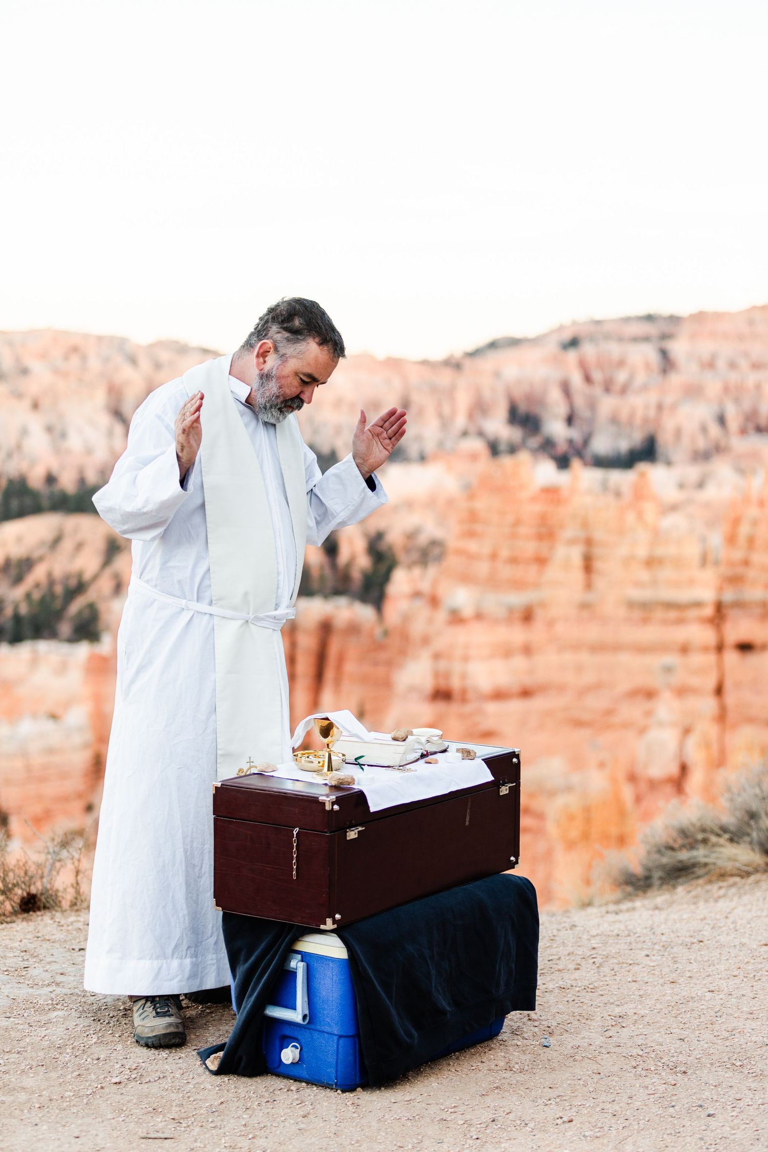 A man in a white robe stands next to a suitcase and a small table in a desert landscape.