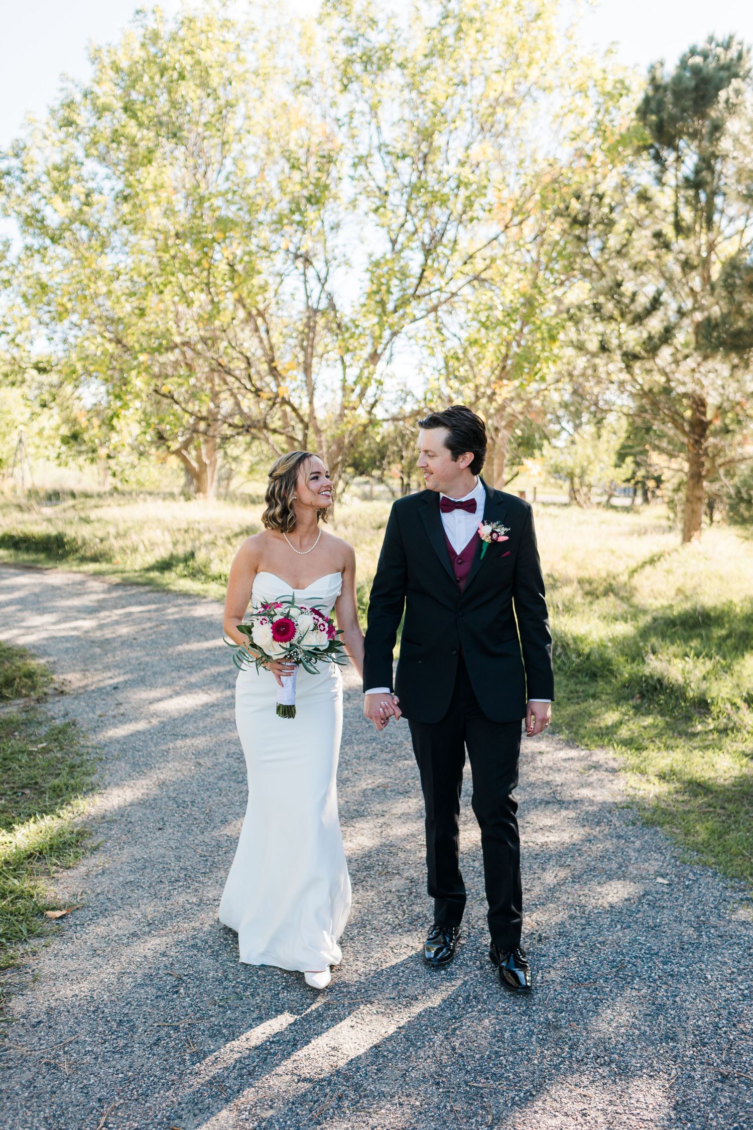 Bride and groom walk hand-in-hand on a gravel path surrounded by trees, she in a white gown, he in a black tuxedo.