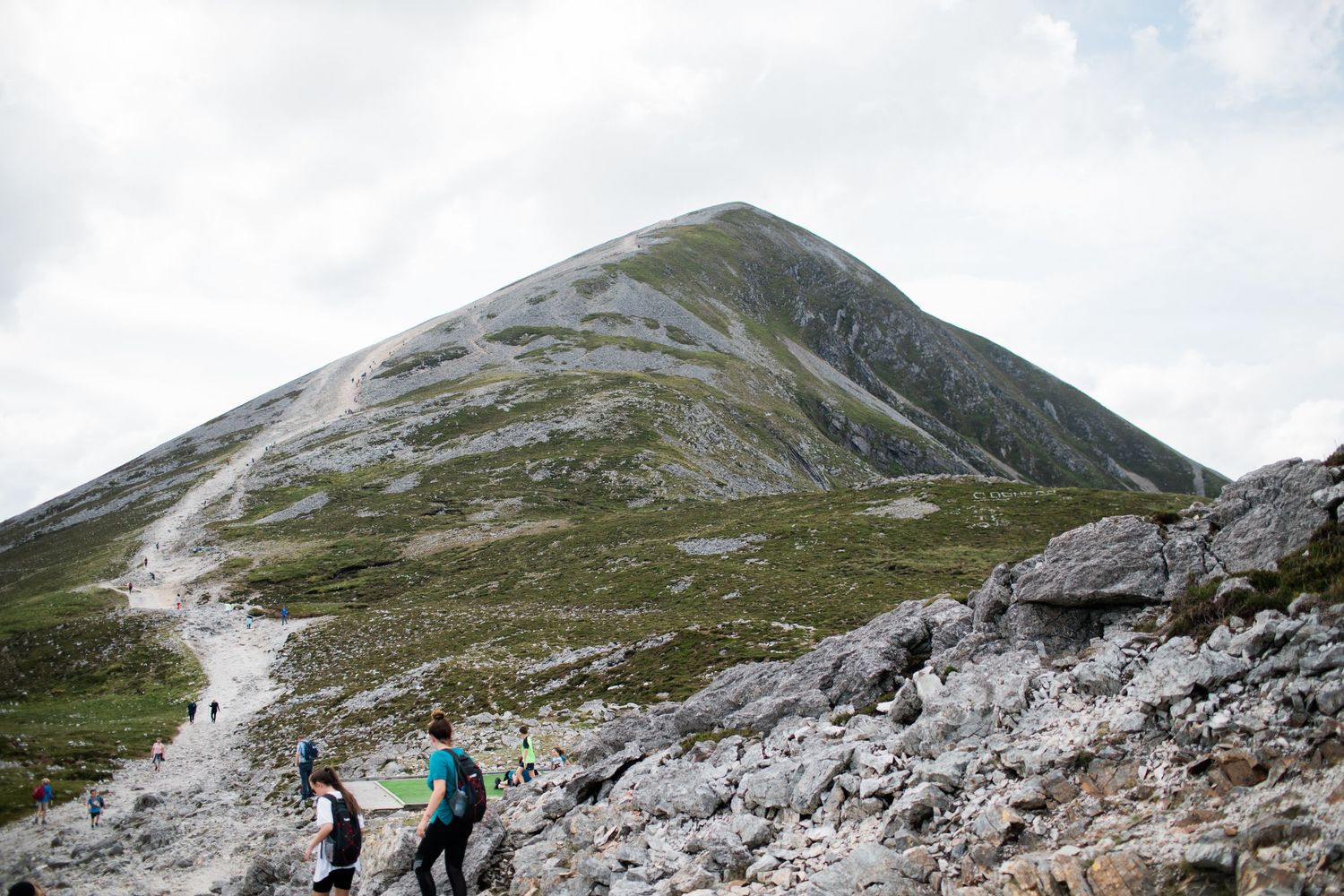 Hikers ascending a rocky mountain trail toward a green pyramid-shaped peak under cloudy skies.