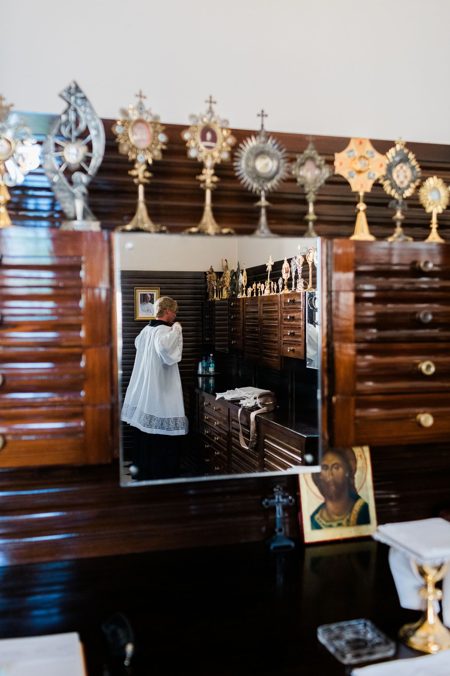 Ornate wooden Catholic sacristy with golden monstrances and a priest reflected in a mirror.