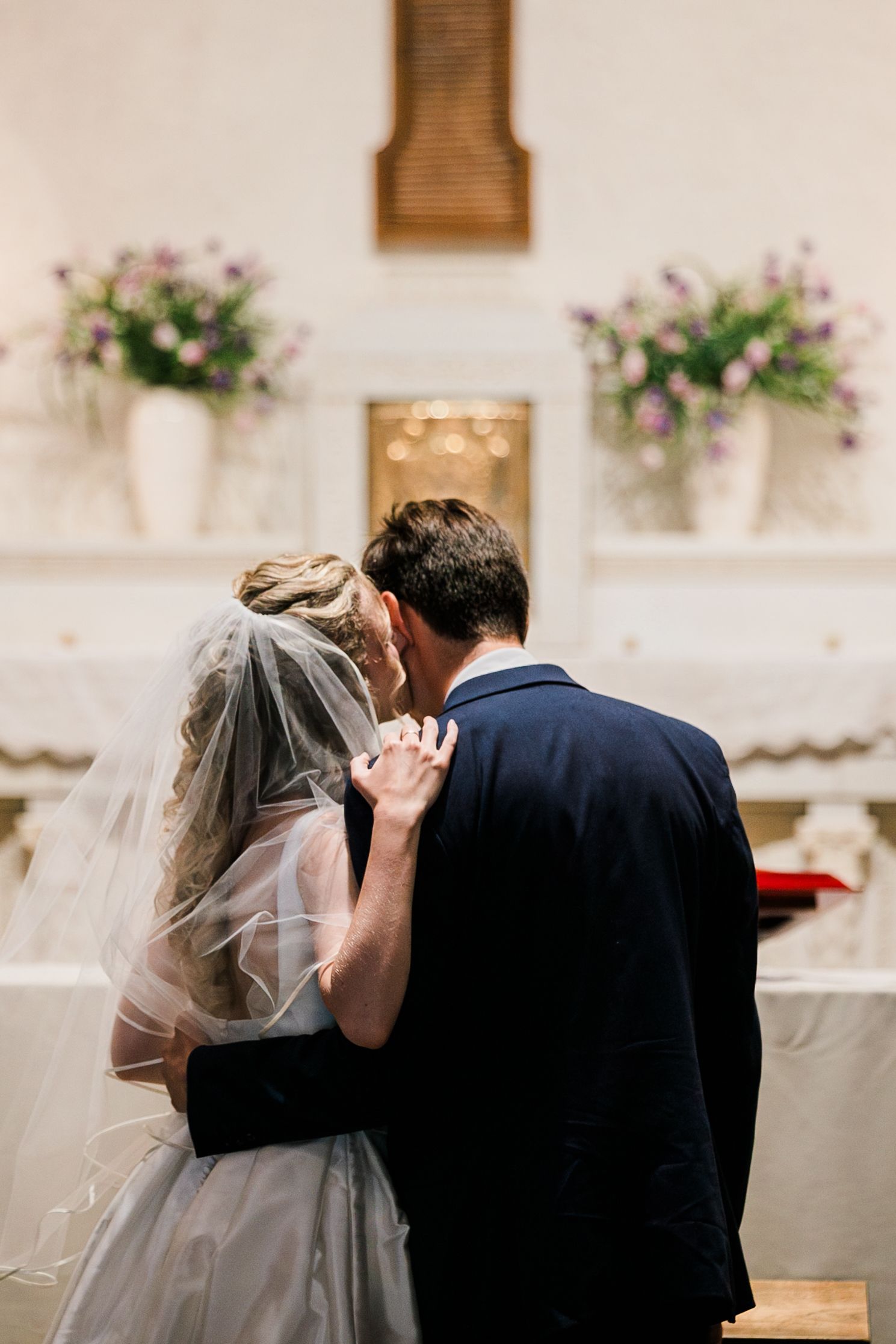 Bride and groom share first kiss at church altar, surrounded by floral arrangements during wedding ceremony.