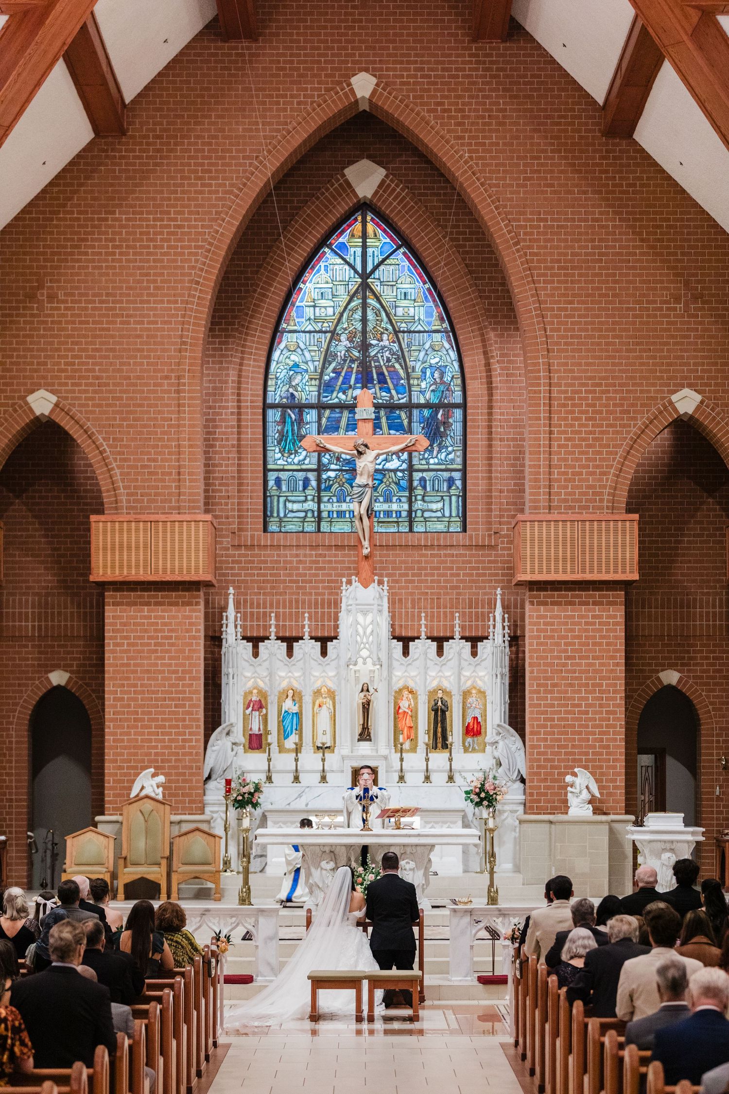 Couple kneeling at church altar during Catholic wedding ceremony with stained glass window and crucifix.