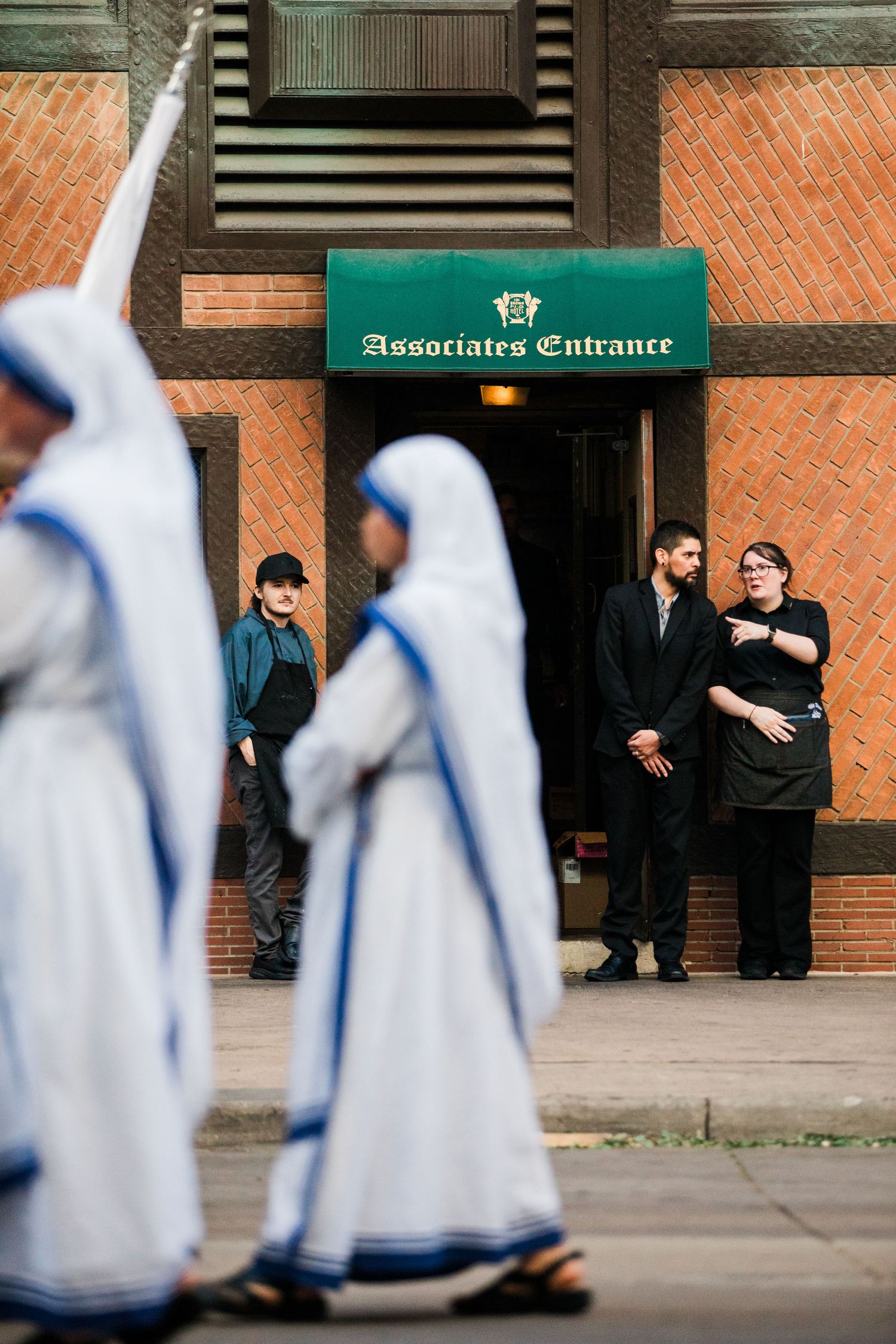 Catholic nuns in white habits with blue trim walk past Associates Entrance as onlookers watch the religious procession.