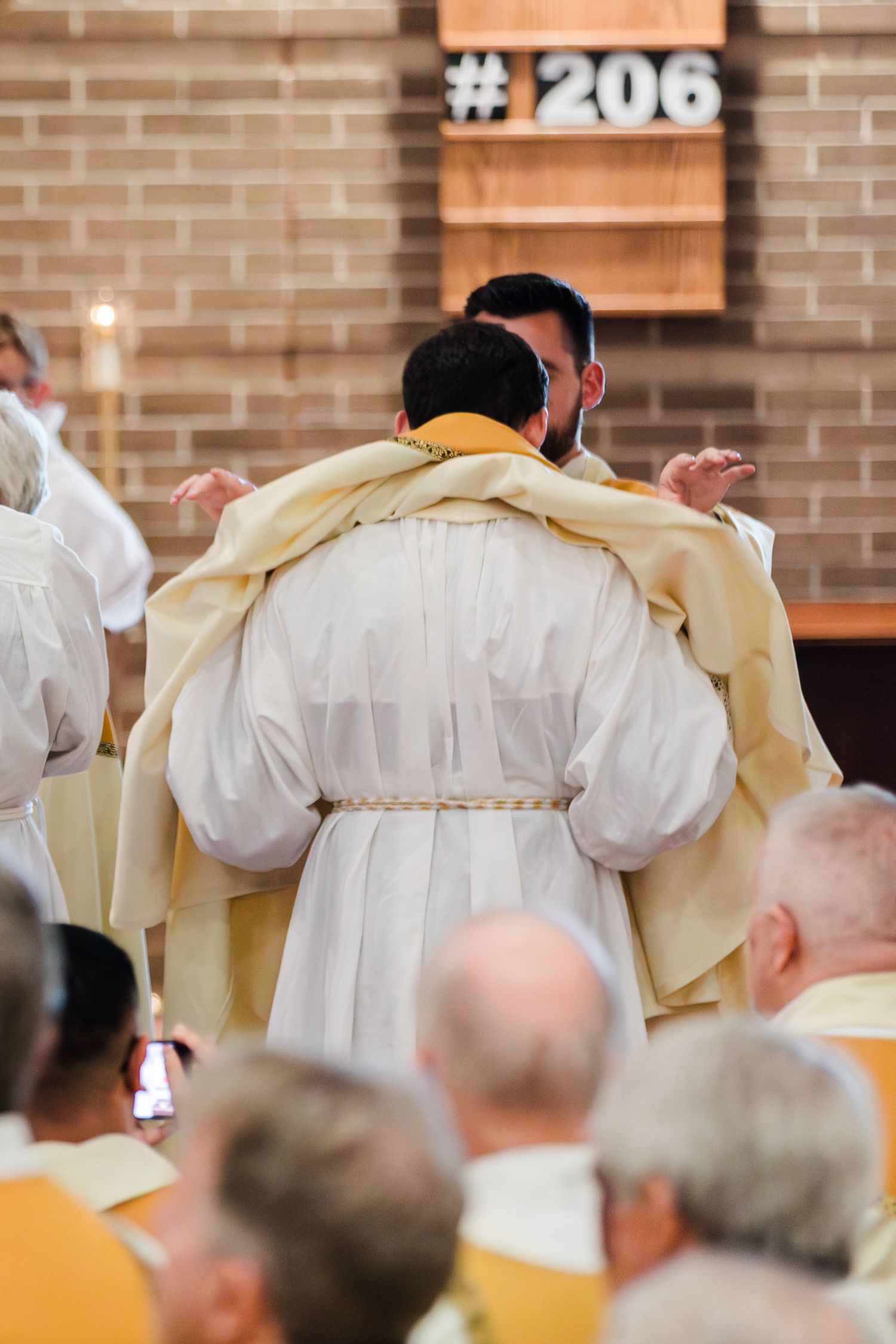 Catholic priest in white vestments bowing during a religious ceremony inside a brick church with congregation seated.