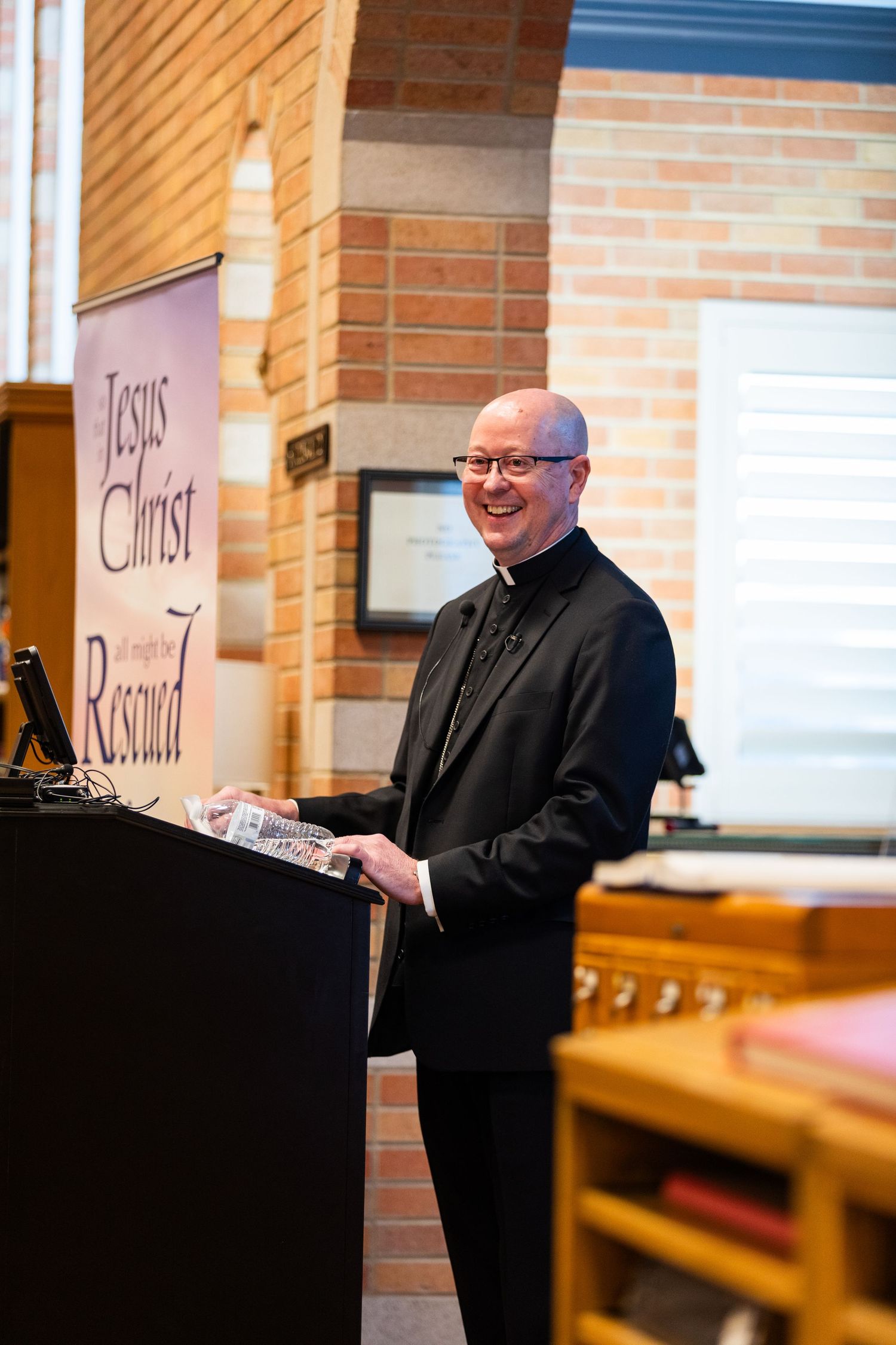 Smiling bald man in black clergy attire stands at podium in church with Jesus Christ banner behind him.