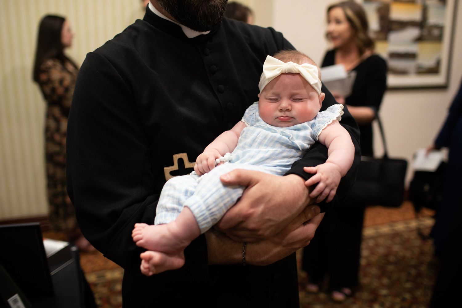 A sleeping infant in white baptismal clothing being held during a church ceremony.