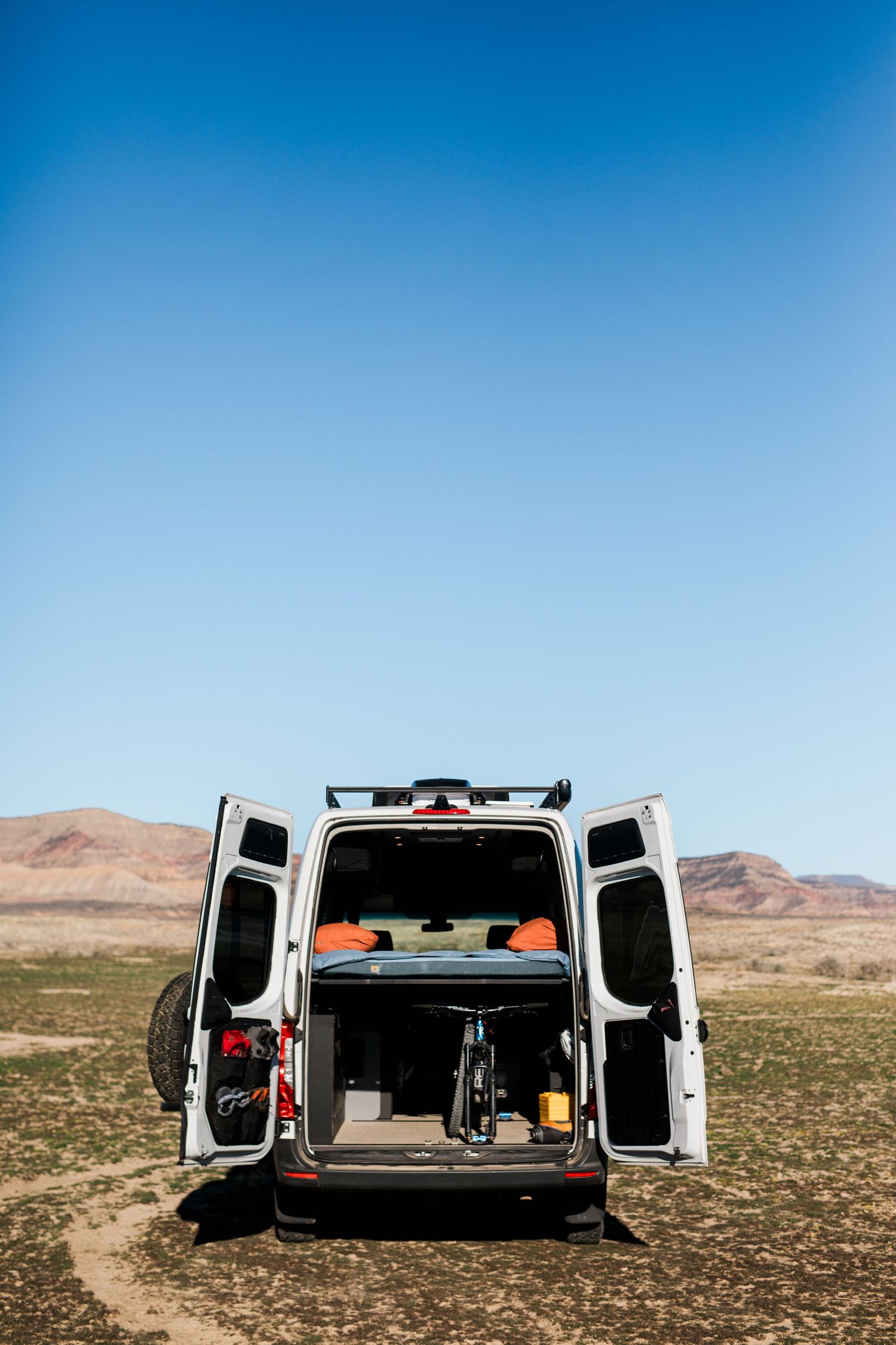 A white van with open back doors parked in a desert landscape with mountains visible in the background.
