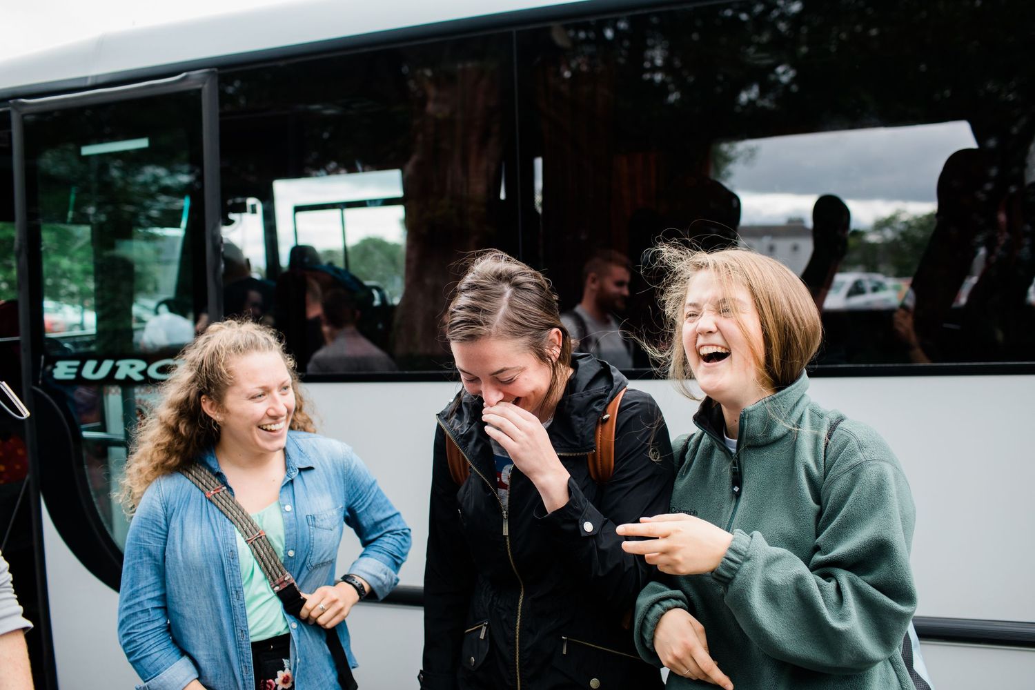 Three young women laughing joyfully together outside a tour bus on a cloudy day.
