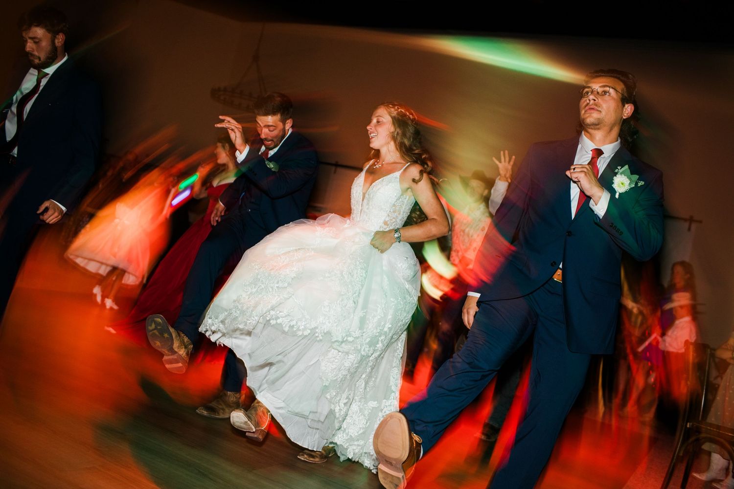 Bride and groom dancing energetically with guests at a vibrant wedding reception under colorful lights.