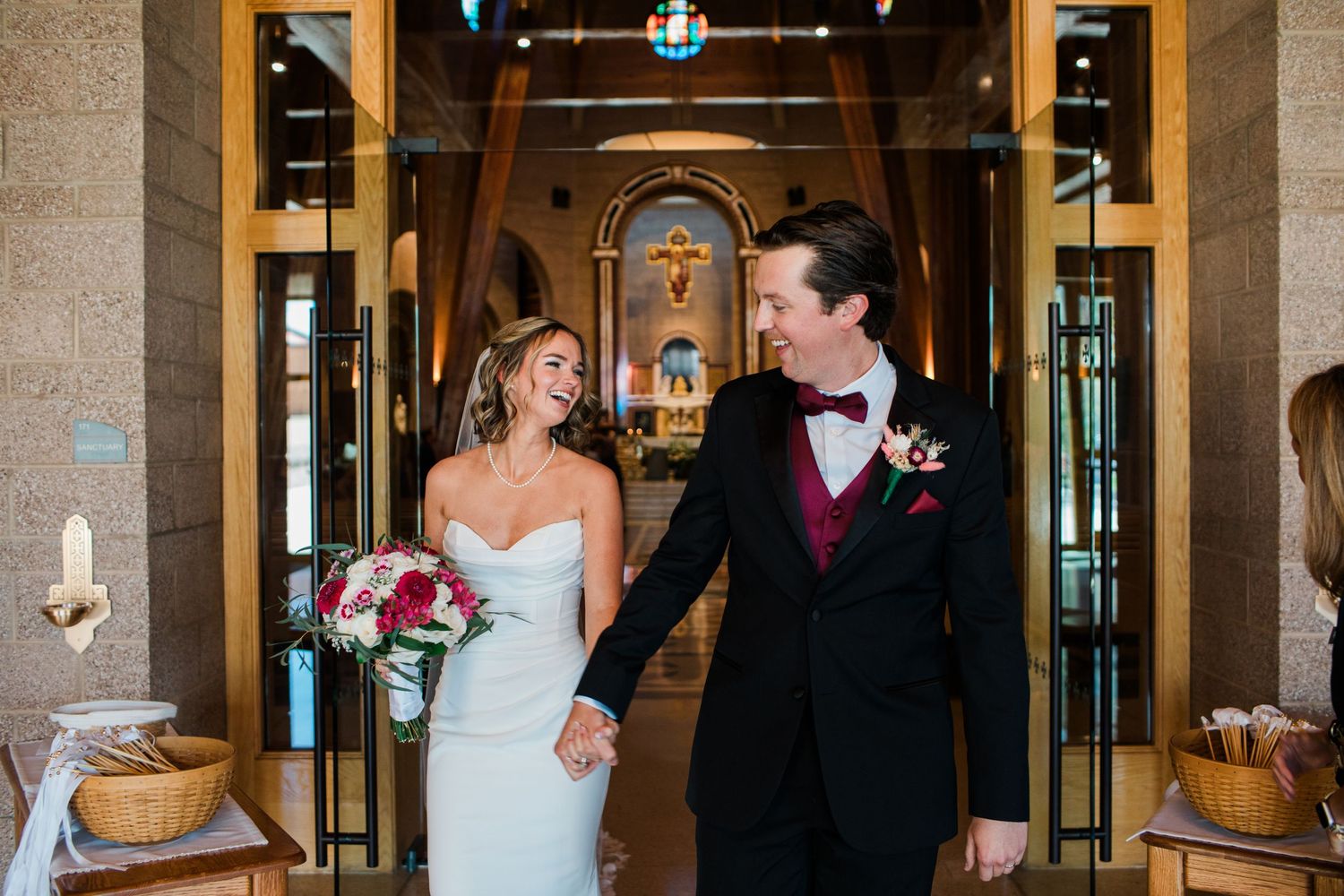Bride and groom holding hands smiling at each other in a church doorway, bride holding red bouquet.