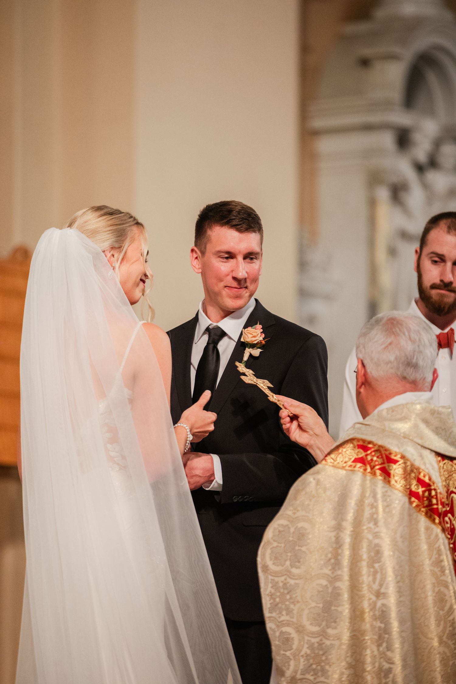 Groom smiling tenderly at bride during ring exchange with priest at elegant church wedding.