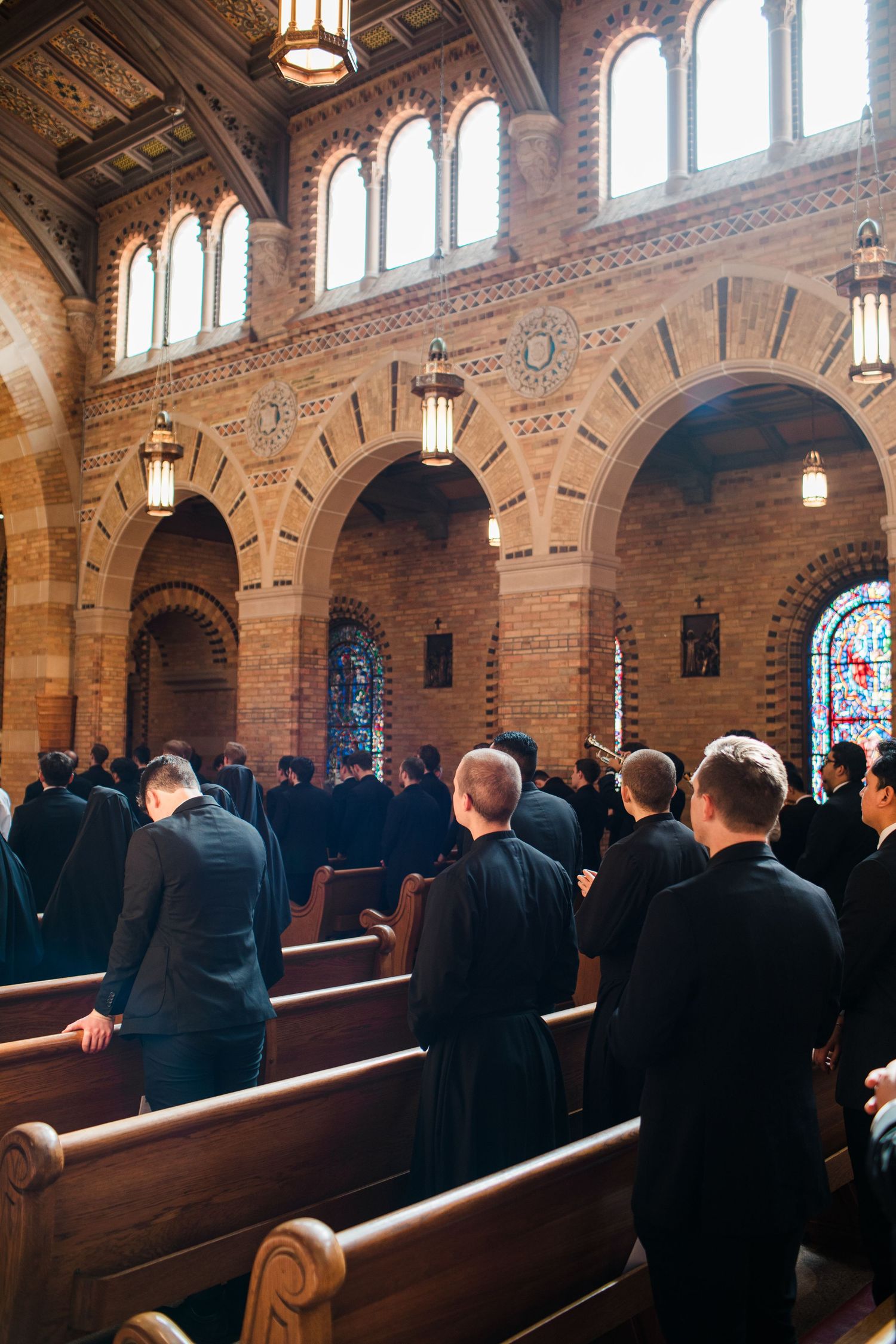 Clergy and congregation stand in prayer inside a grand brick Catholic church with arched windows and stained glass.