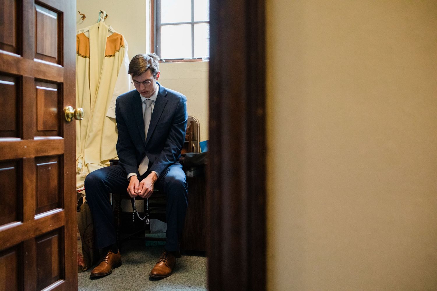 A young man in a navy suit sits pensively in a small room, hands clasped, head bowed, viewed through a doorway.