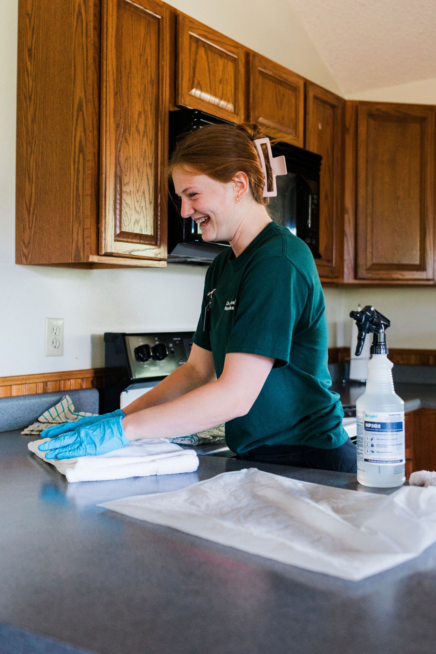 Smiling woman in green scrubs and blue gloves cleaning a kitchen counter with a spray bottle nearby.