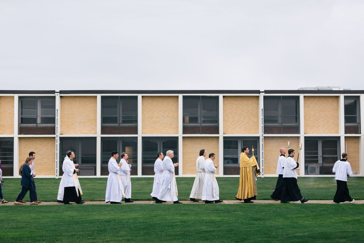 People in formal attire stand in line along a building exterior with grass and windows in the background.