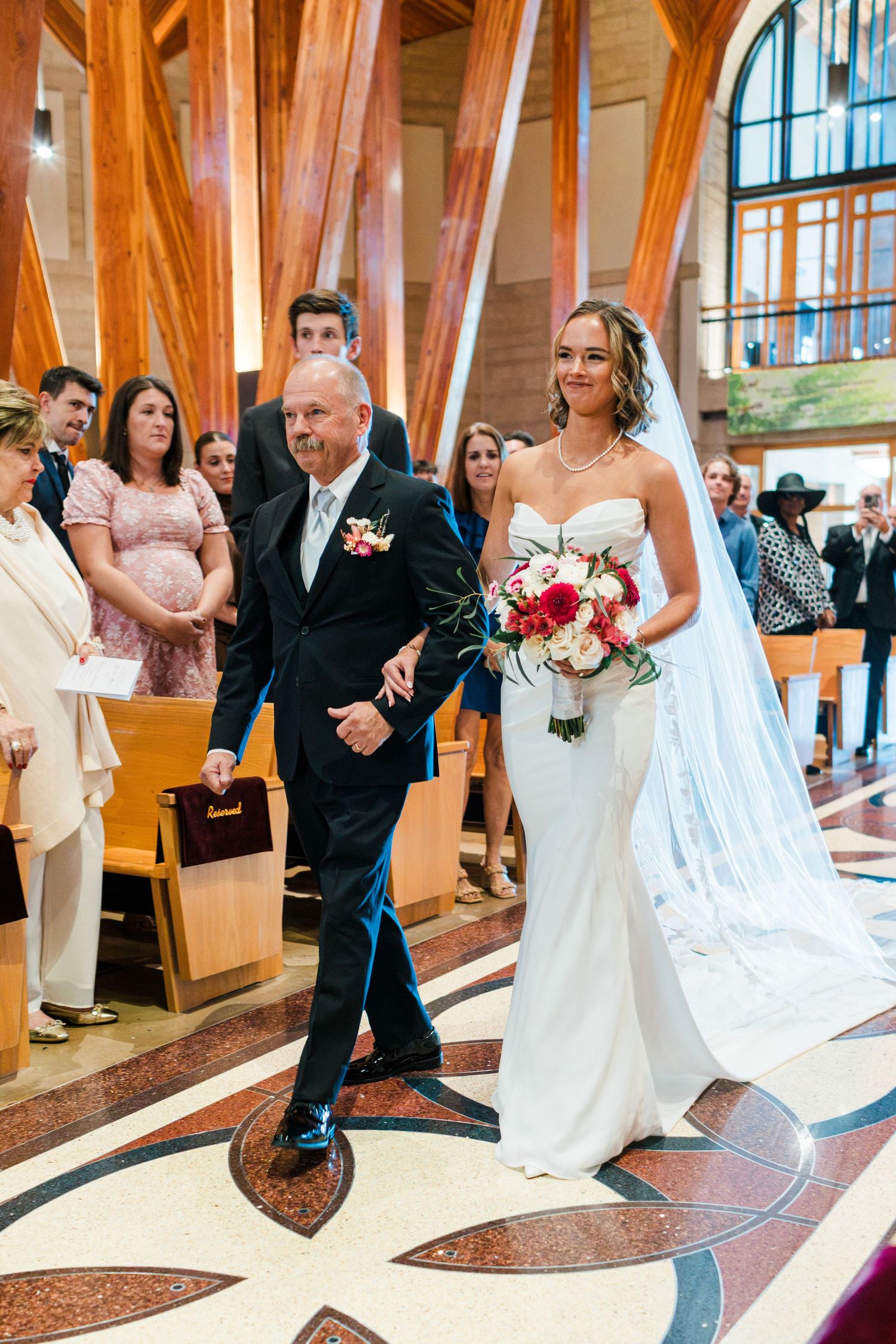 Bride walks down church aisle with escort holding colorful bouquet during wedding ceremony.