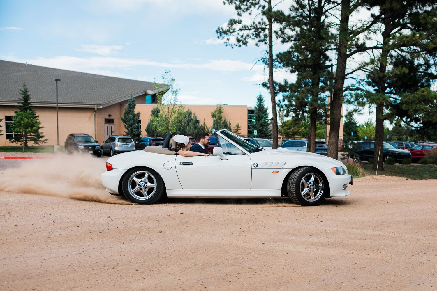 White convertible sports car drifting on a dirt lot, kicking up dust clouds near trees and a building.
