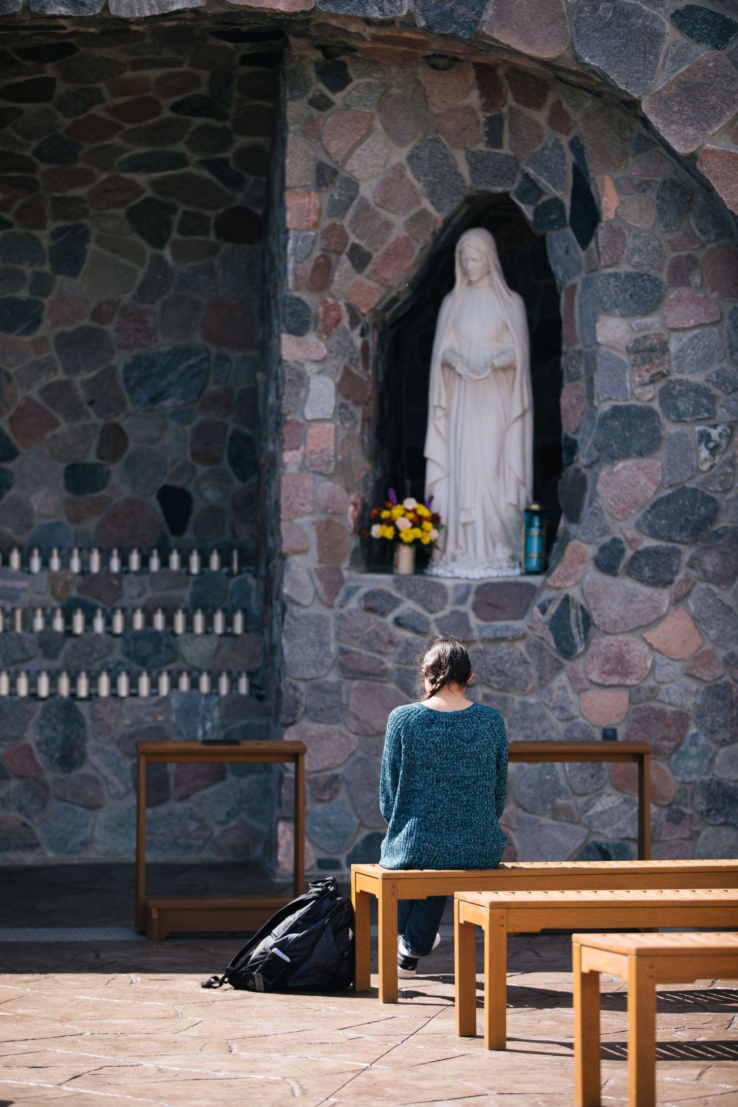 Person sits quietly on wooden bench in stone church alcove with religious statue in contemplative meditation.