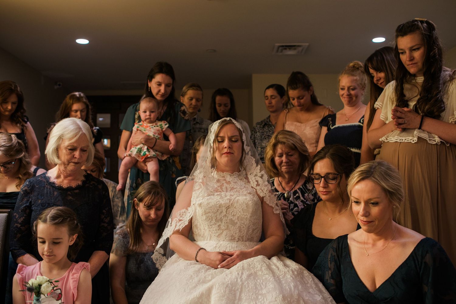 Bride in white lace gown and veil surrounded by bridesmaids and family members in a warmly lit room.