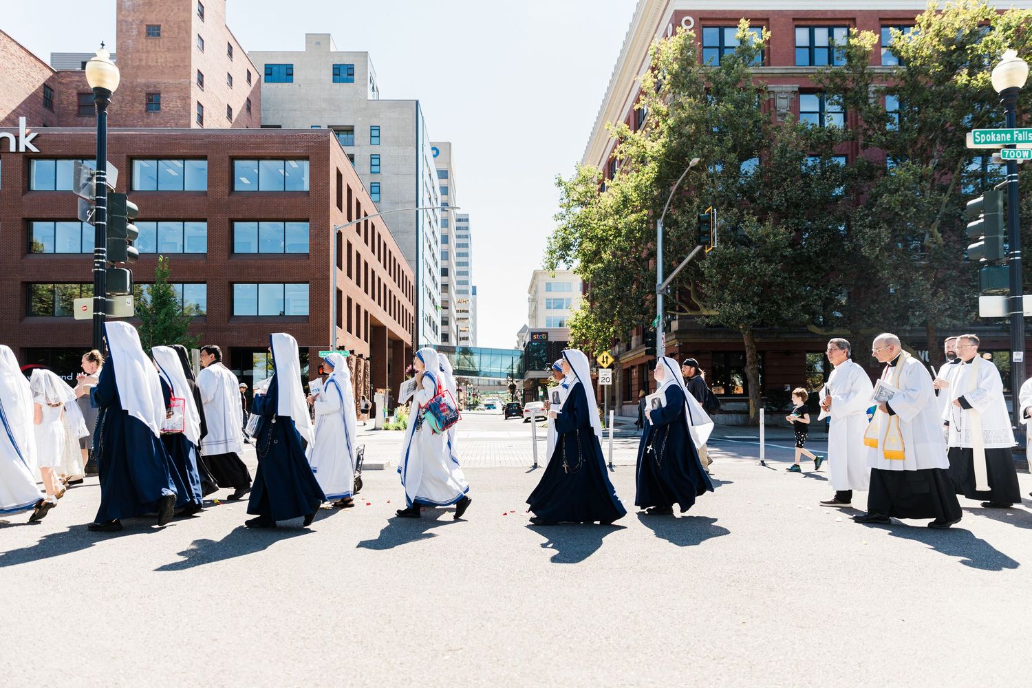 Long panoramic view of people walking along an urban sidewalk with brick buildings in background.
