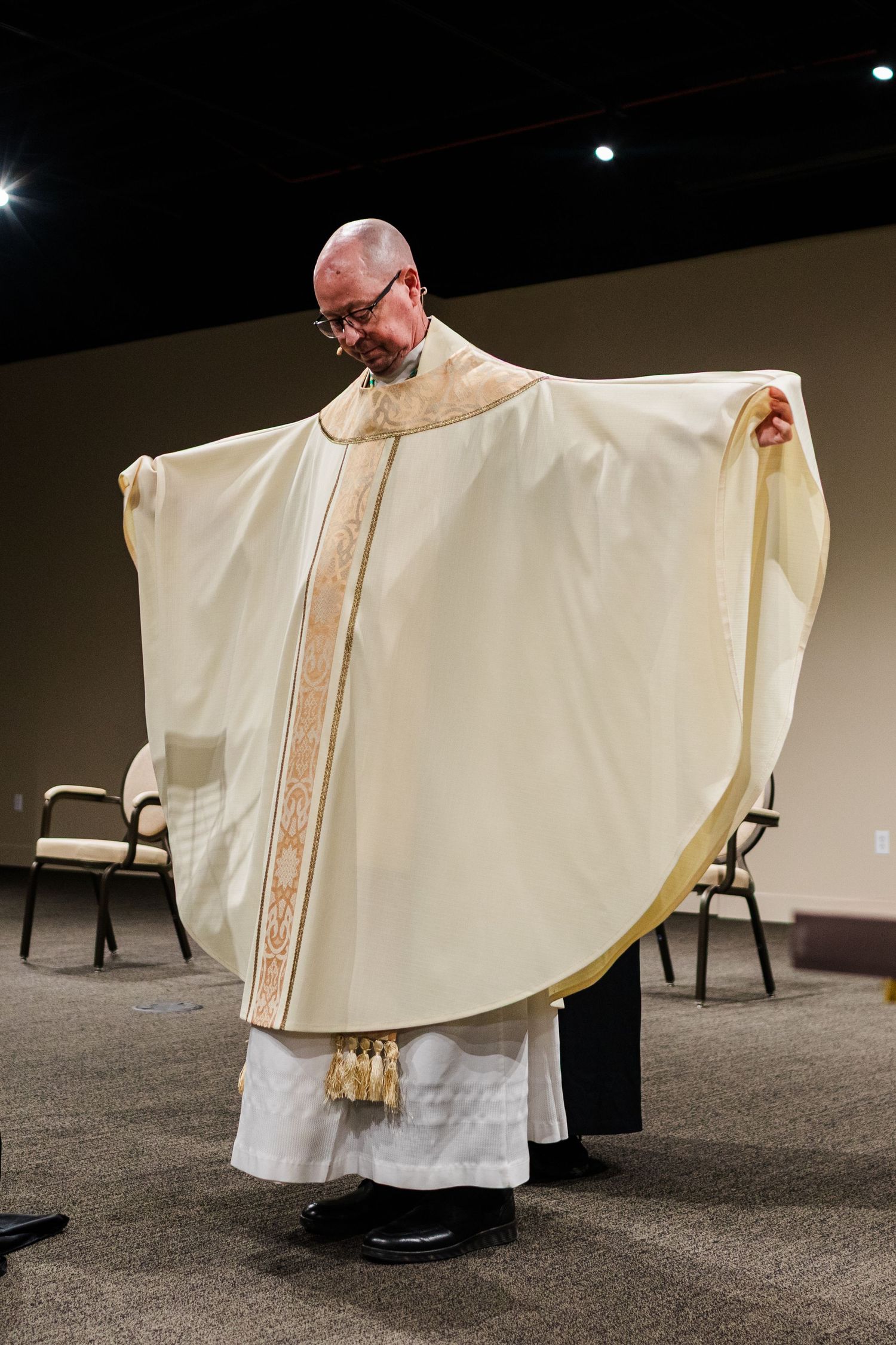 Clergy member wearing white ceremonial chasuble with gold trim stands displaying the garment during religious service.
