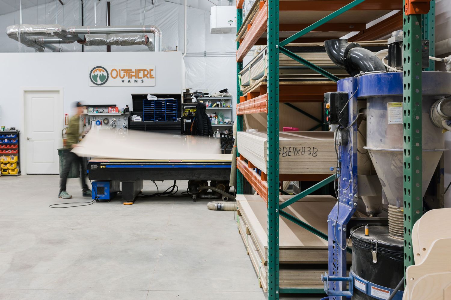 Inside a warehouse, a person works at a large machine with industrial storage shelves visible in the foreground.