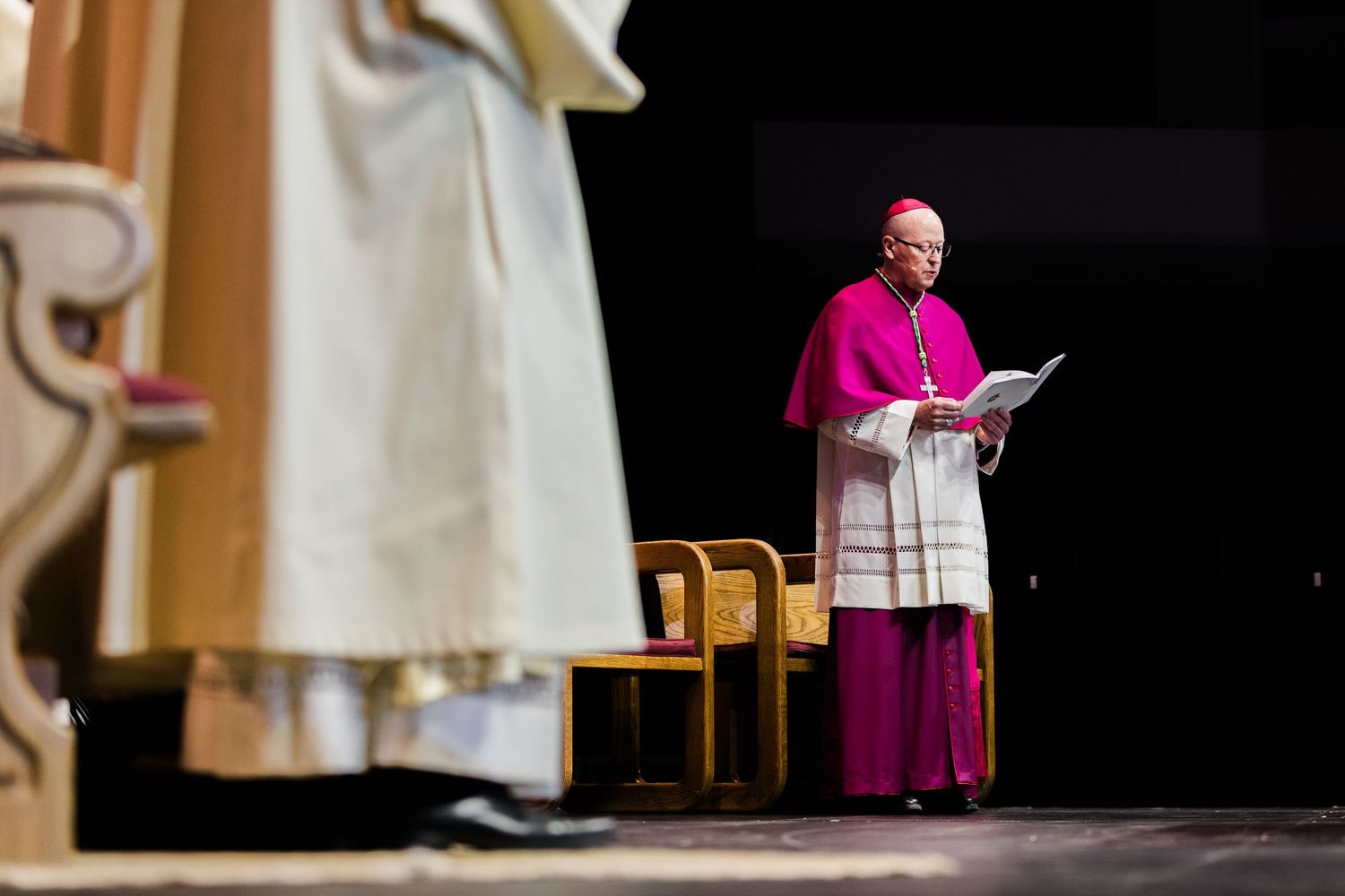 A Catholic bishop in purple vestments reads from papers during a formal religious ceremony indoors.