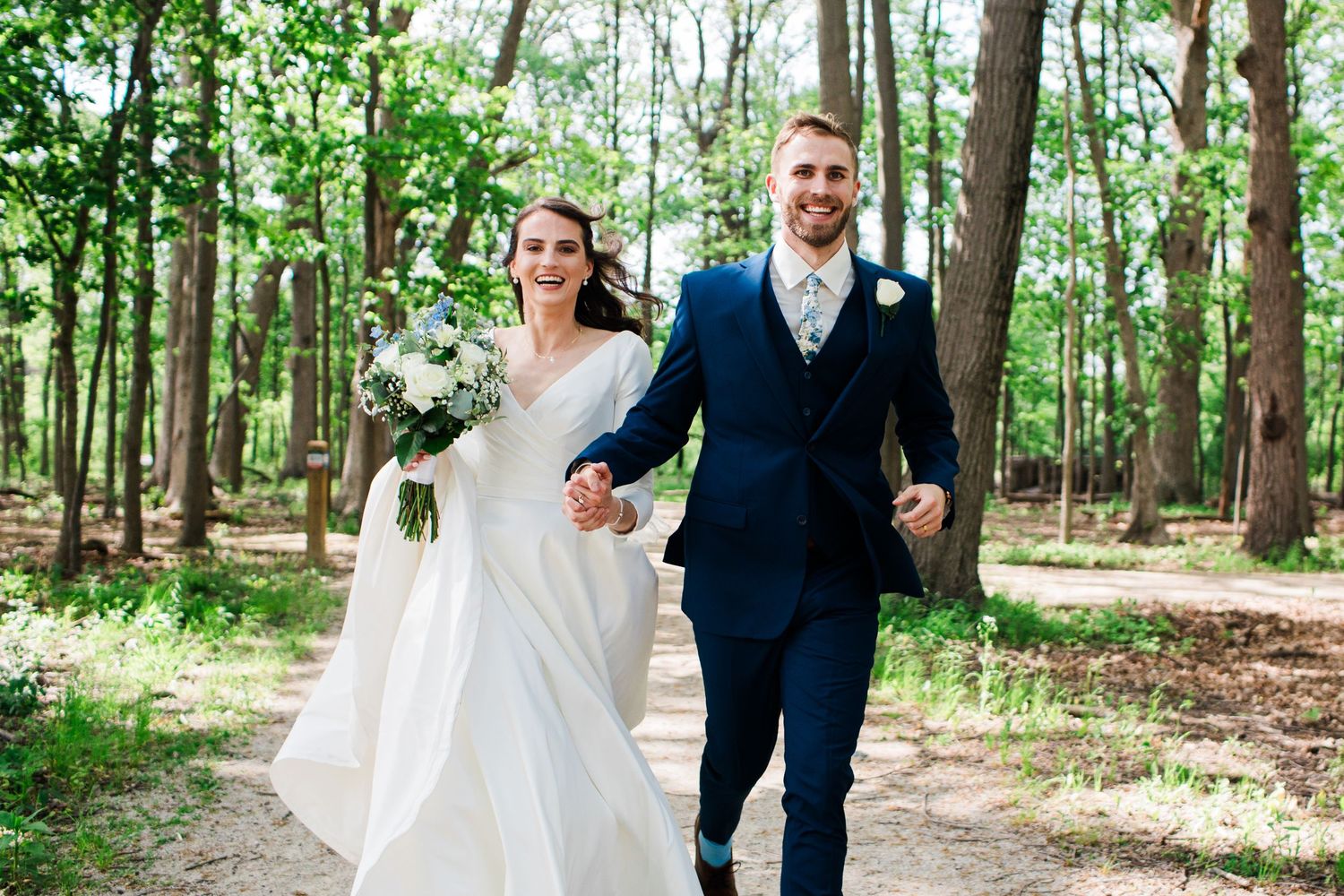 Happy bride and groom walking hand-in-hand through a sunlit forest on their wedding day.