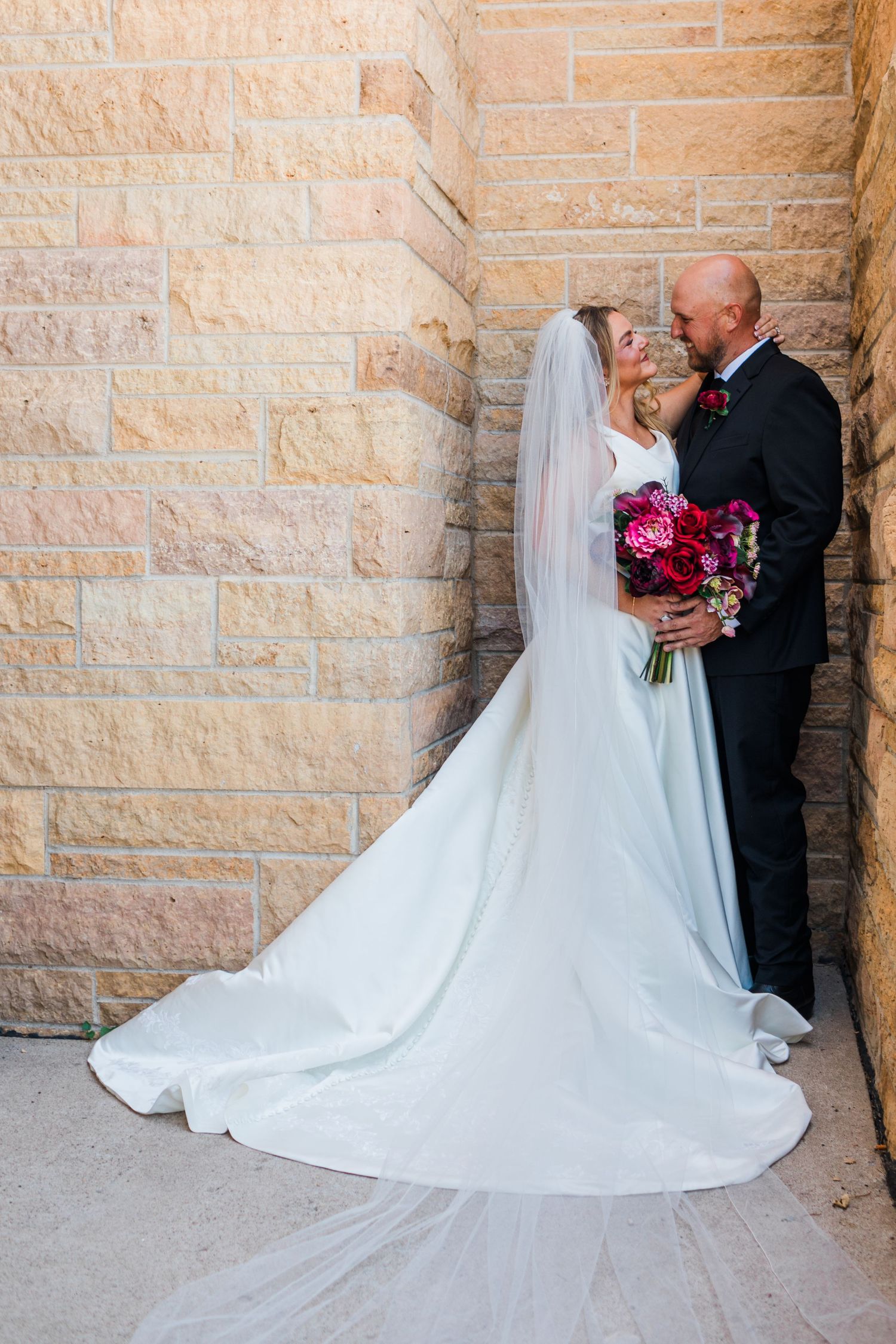 Bride in white gown with veil and groom in black suit share a kiss against a stone wall, holding a vibrant pink bouquet.